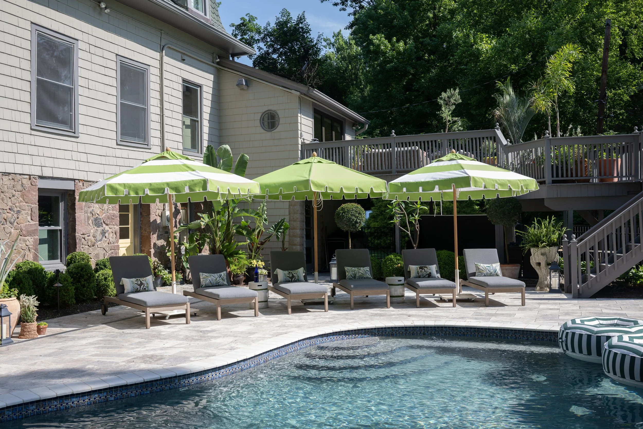 Residential backyard with a swimming pool, green patio umbrellas, lounge chairs with pillows, potted plants, and a deck with stairs. There are trees and a house in the background.