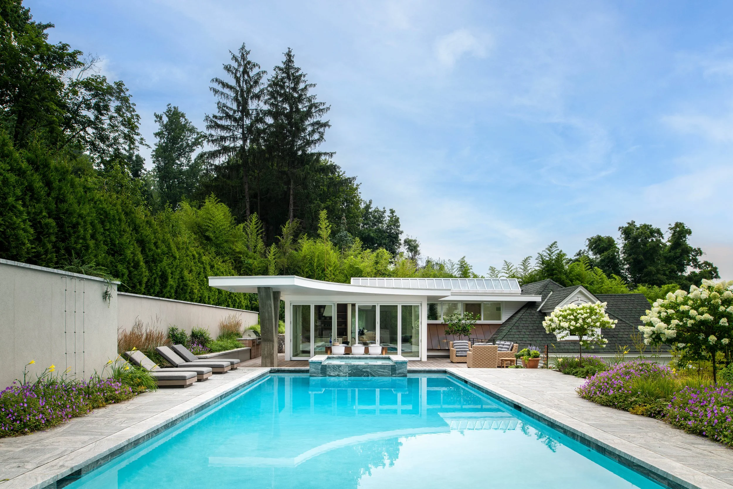 A modern backyard with a swimming pool, surrounded by lush green trees and colorful flowers, and a house with large windows and outdoor seating.