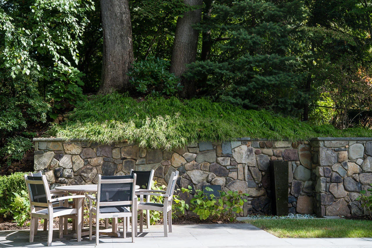 Outdoor garden patio with stone wall, green plants, trees, and a round table with six chairs.