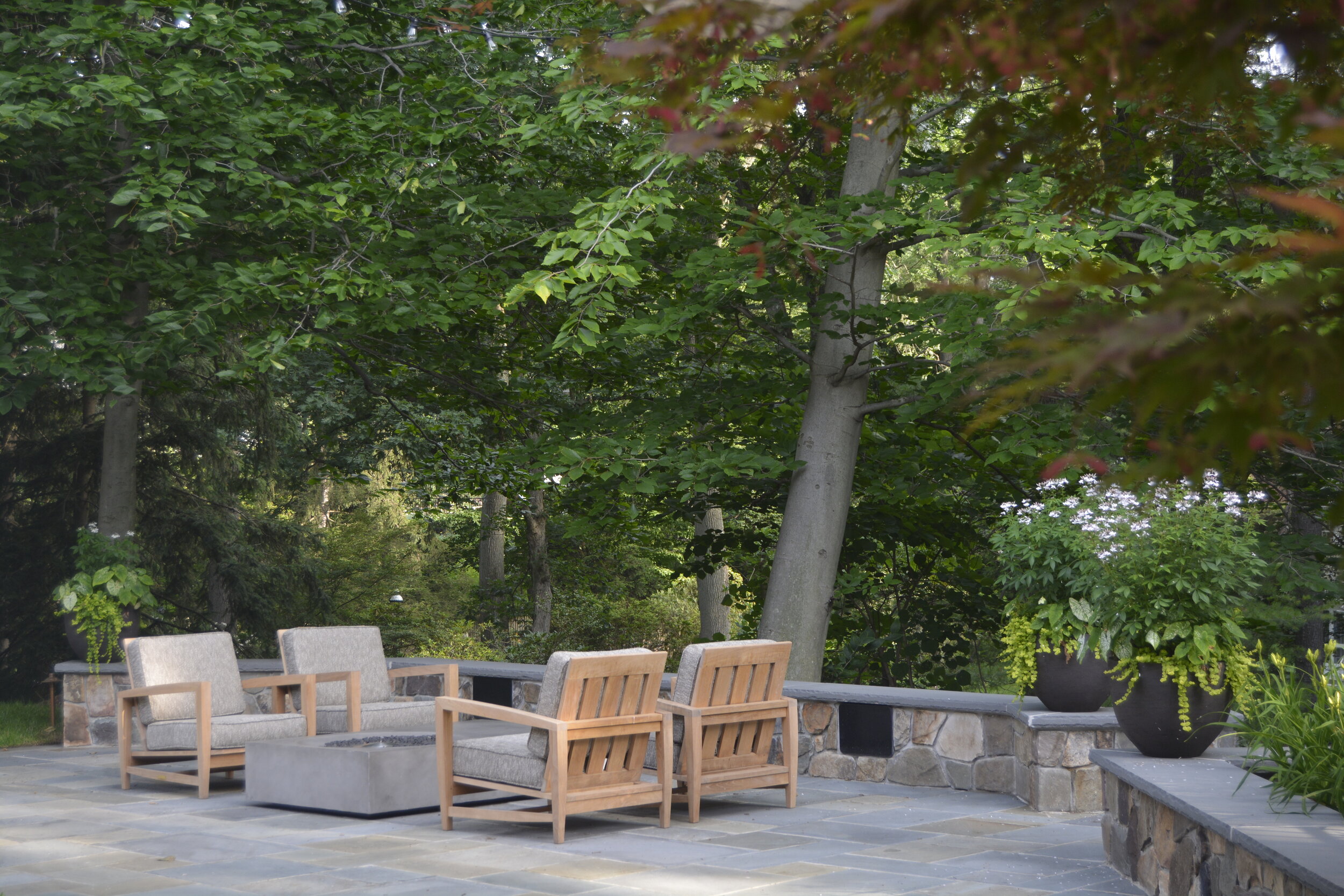 Outdoor patio with gray stone flooring, wooden and cushioned chairs, stone wall with large black planters, green trees in the background.
