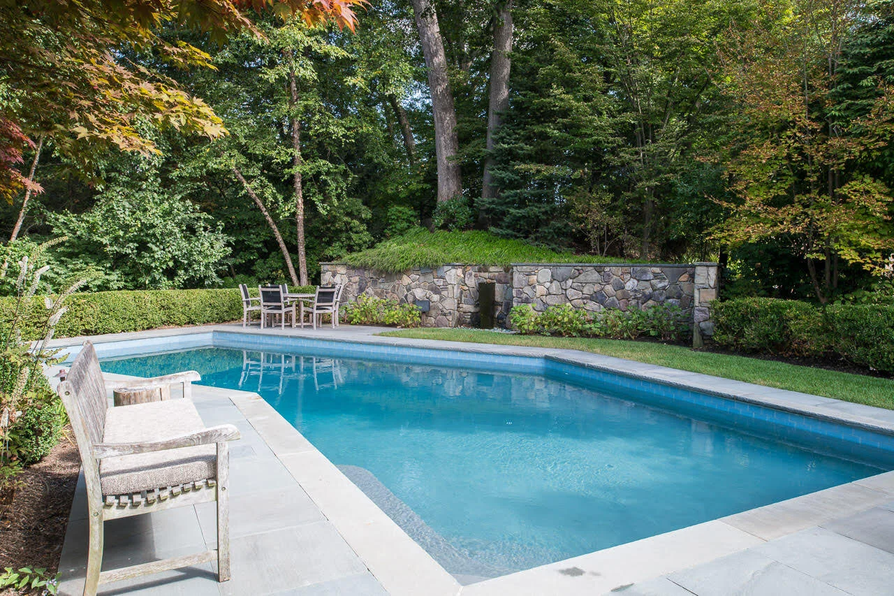 A backyard swimming pool surrounded by trees and shrubs with a stone wall in the background, outdoor furniture including a bench and a table with chairs, and a grassy area.