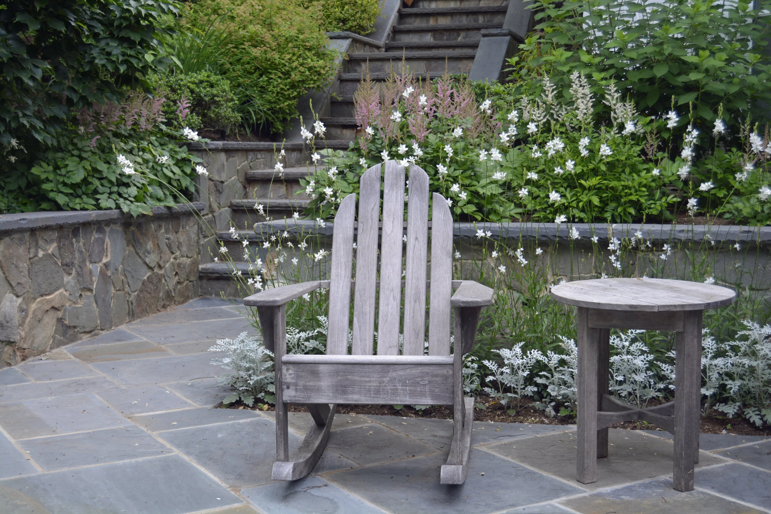 A weathered Adirondack rocking chair and a small matching side table sit on a stone patio surrounded by flowering plants and greenery.