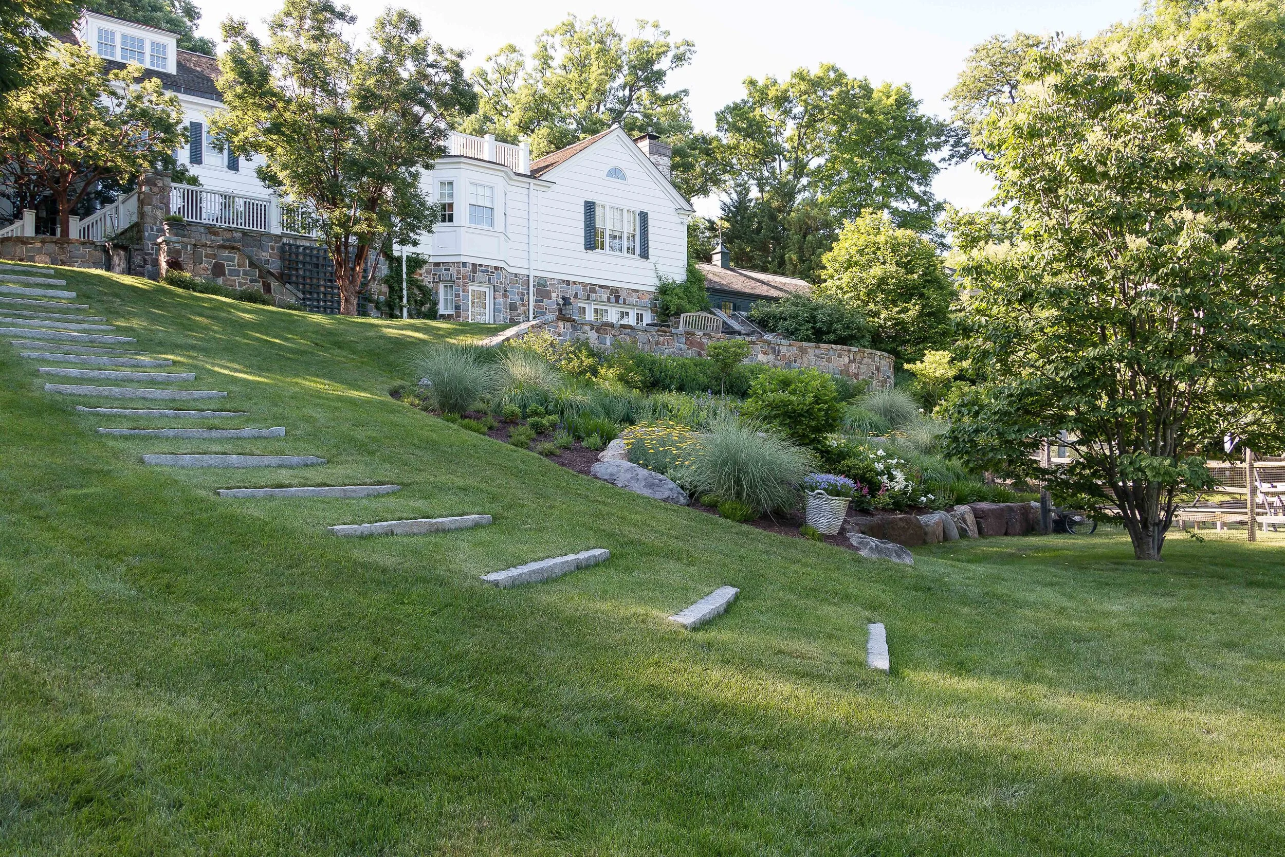 Tilted view of a lush green backyard with stone steps on the grassy slope, landscaping with bushes, flowers, and trees. A white house with black shutters and a stone foundation is visible in the background.
