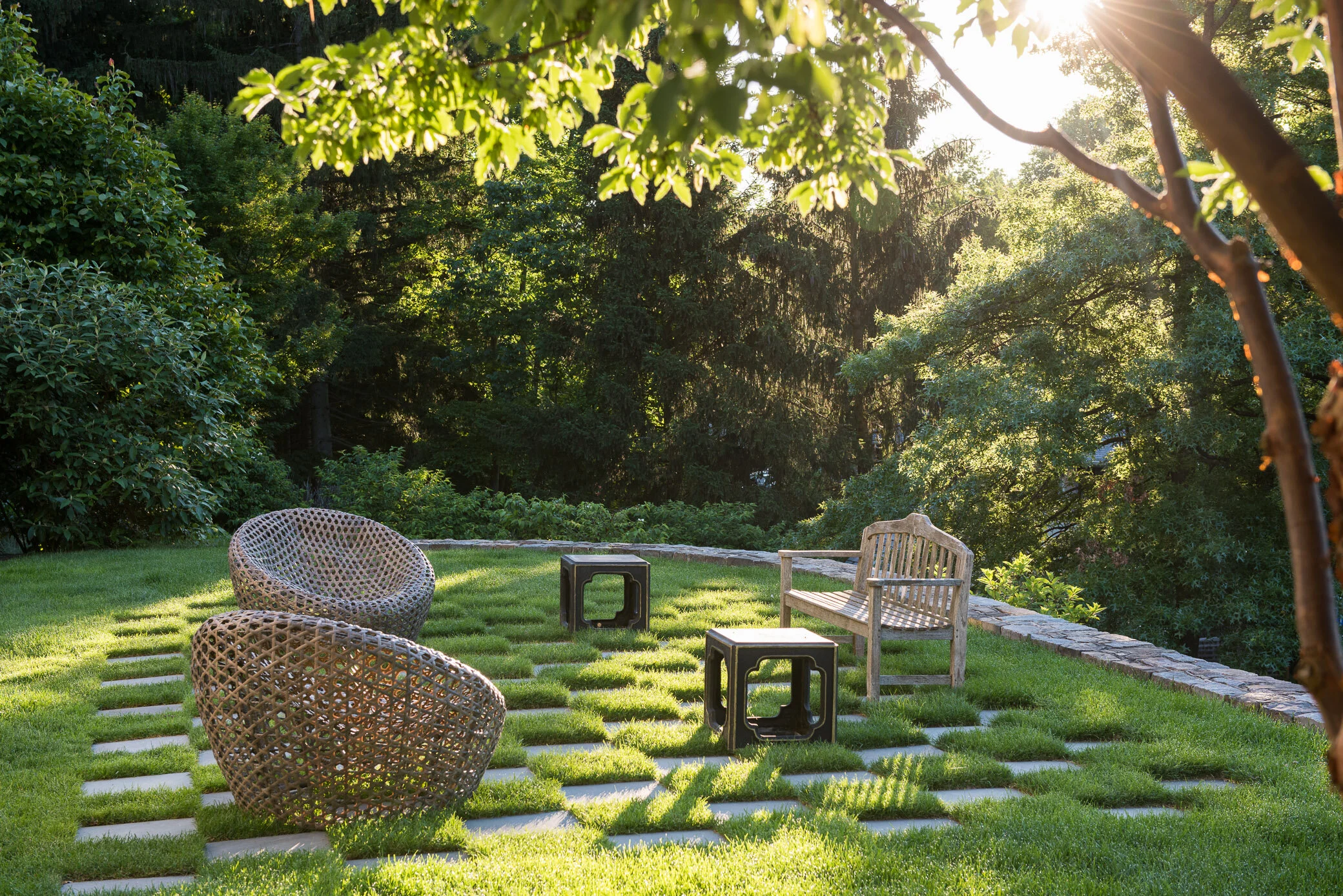 A backyard with green grass and trees, illuminated by sunlight, with a wooden bench, two wicker chairs, and two small black tables arranged on a stone and grass patio.