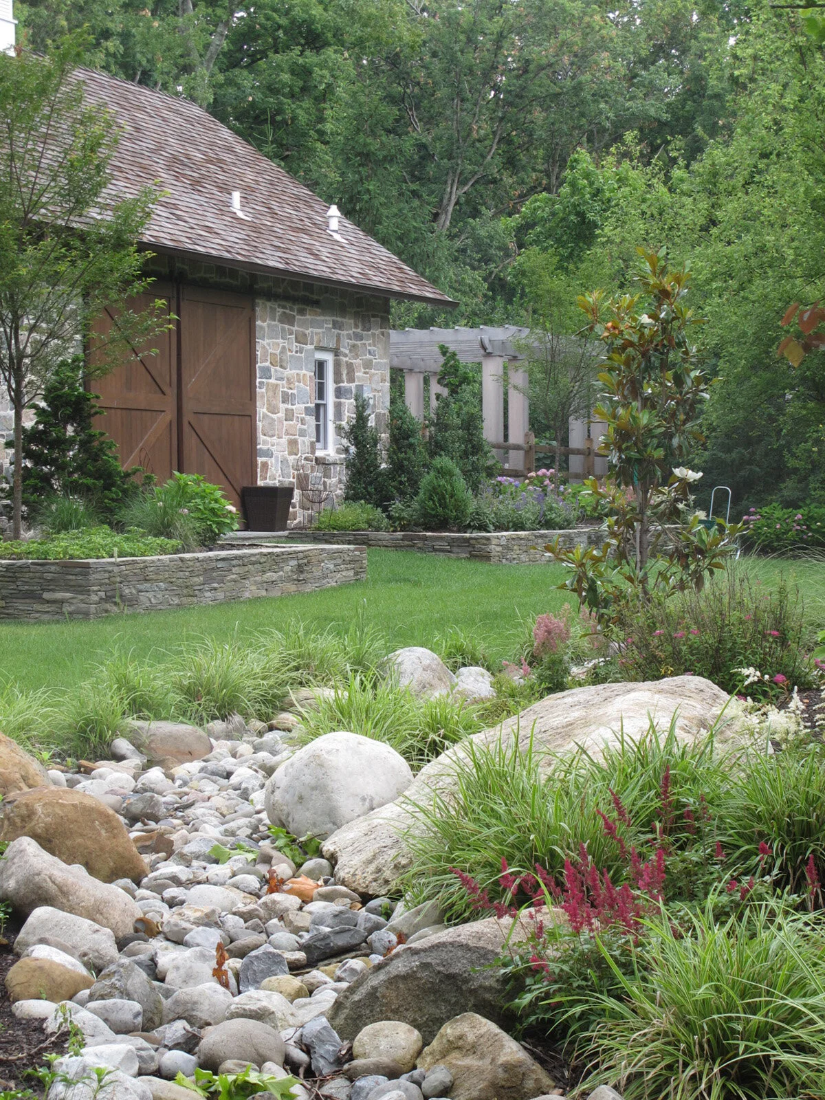 A landscaped backyard garden with colorful flower beds, large rocks, and trimmed grass, with a stone barn-style building and lush green trees in the background.