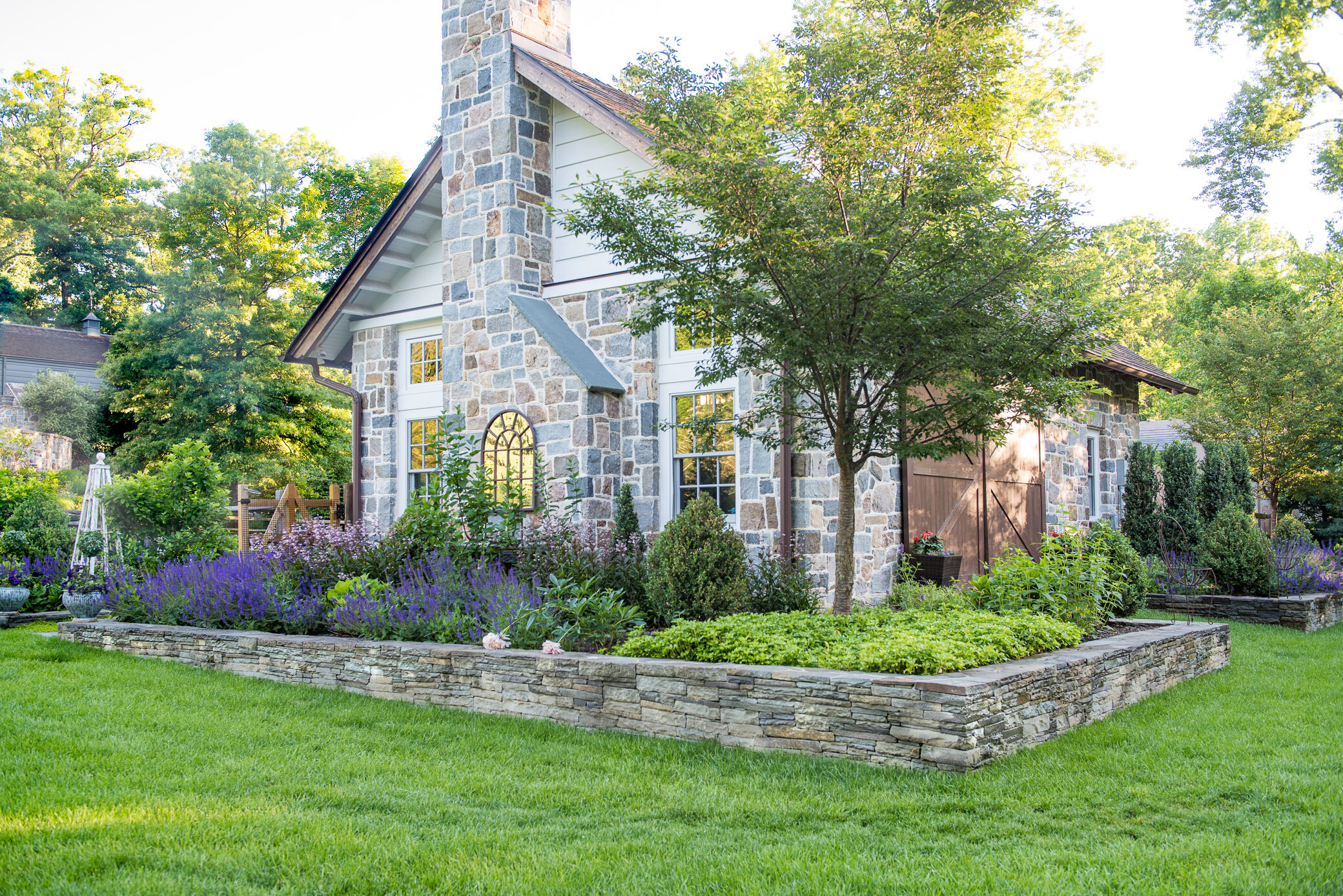 A well-manicured front yard garden with lush green grass, purple flowers, and a small tree in front of a house with stone and white siding, multiple windows, and wooden shutters.