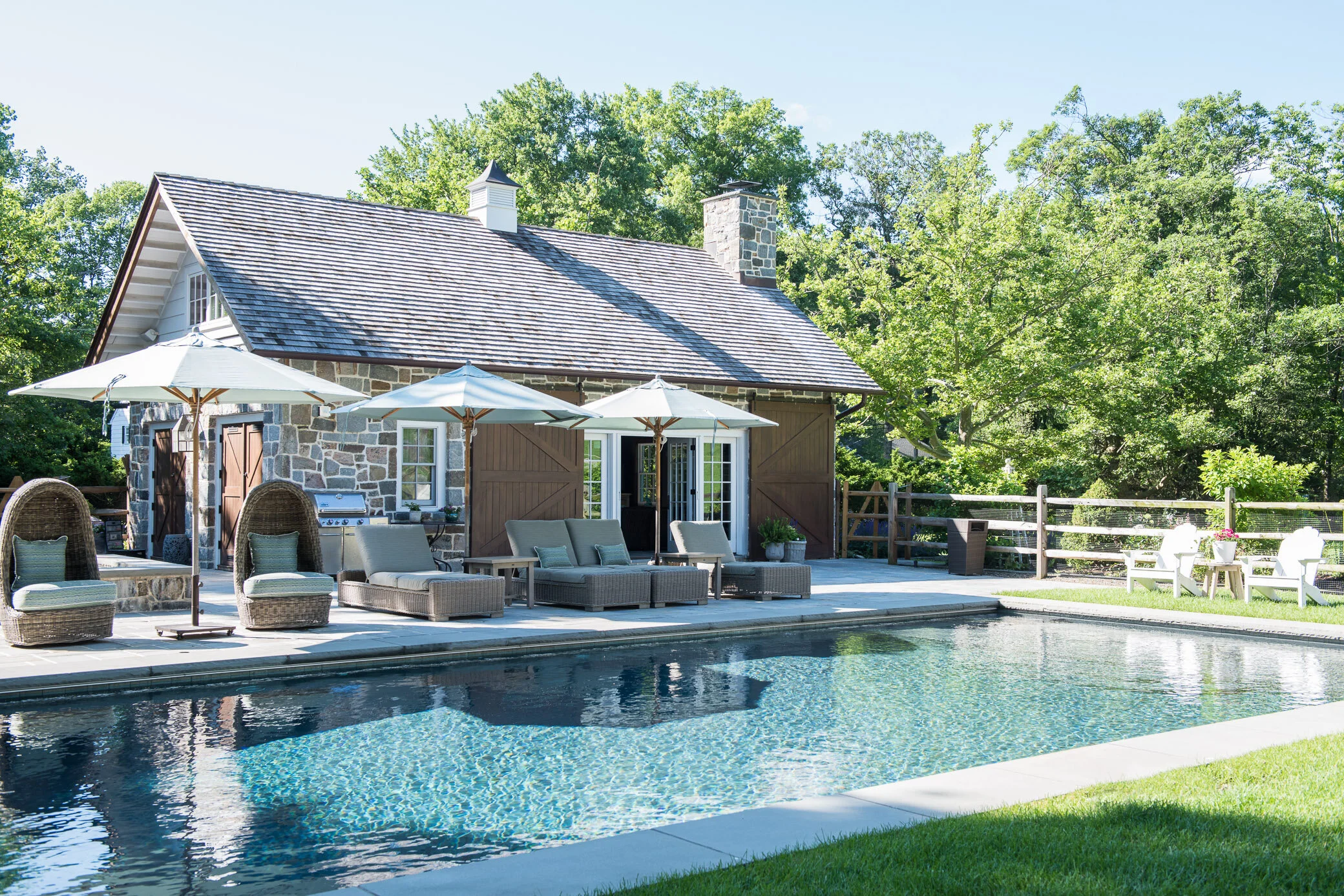 A backyard with a swimming pool, outdoor furniture with umbrellas, a stone house with a shingled roof, and a wooded area in the background.