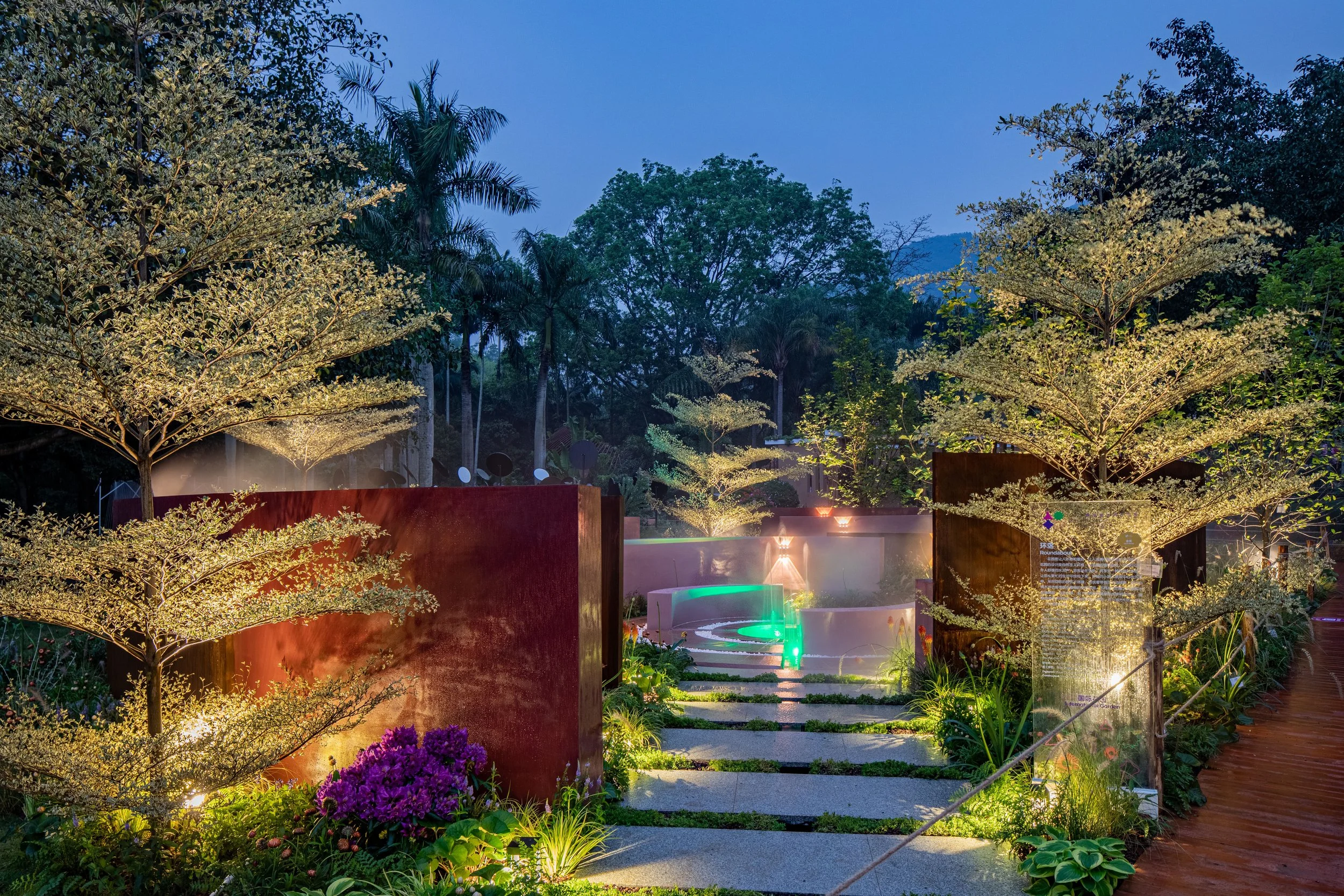 Illuminated garden scene at dusk with trees, pathway, and modern structures.