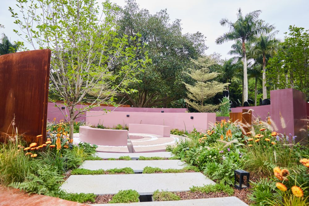 A landscaped garden with a pathway, pink walls, colorful flowers, green bushes, and trees, with a seating area in the background.