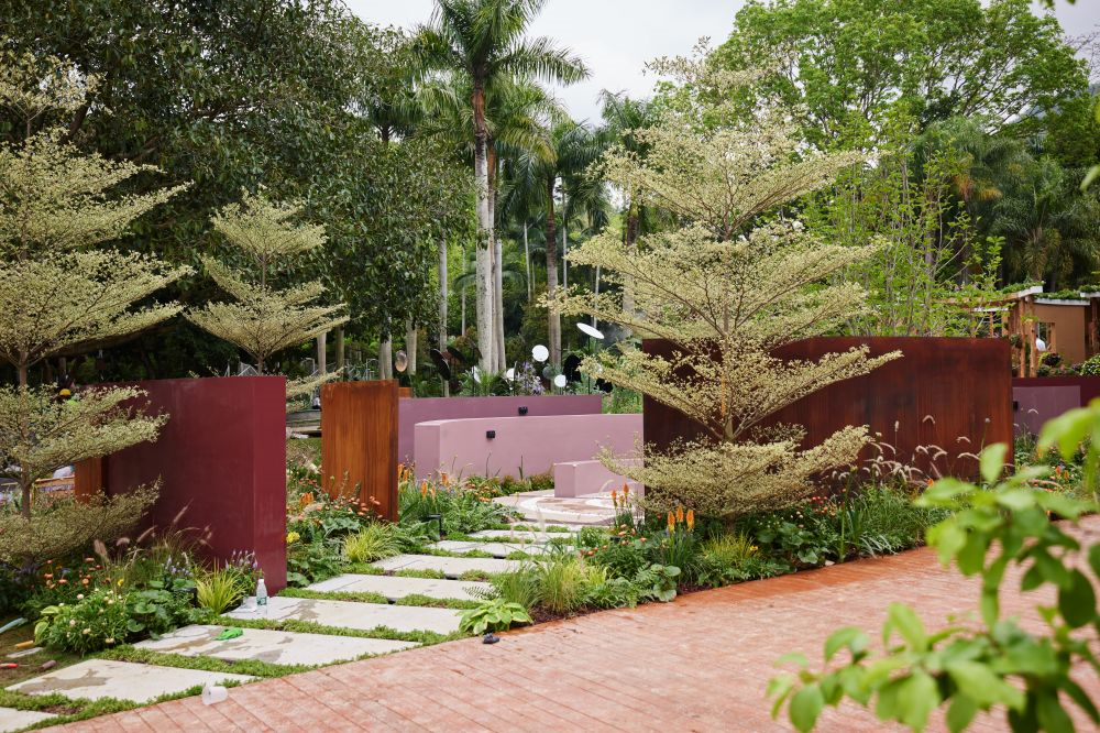 A landscaped garden with stepping stones, colorful flowering plants, small trees, and large decorative metal panels in various earthy colors, surrounded by lush green trees.