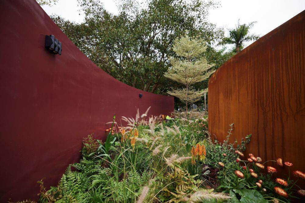 A garden pathway between two modern, curved walls—one red and one wood-colored—surrounded by various plants and flowers, with trees and a cloudy sky in the background.
