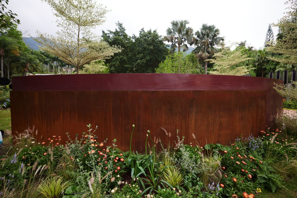 A rust-colored metal fountain with a curved top, surrounded by various plants and flowers in a garden.