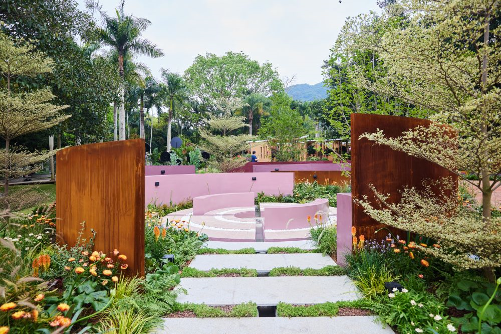 A modern garden with pink and purple concrete structures, surrounded by lush greenery and flowers, and rust-colored metal panels on the sides.