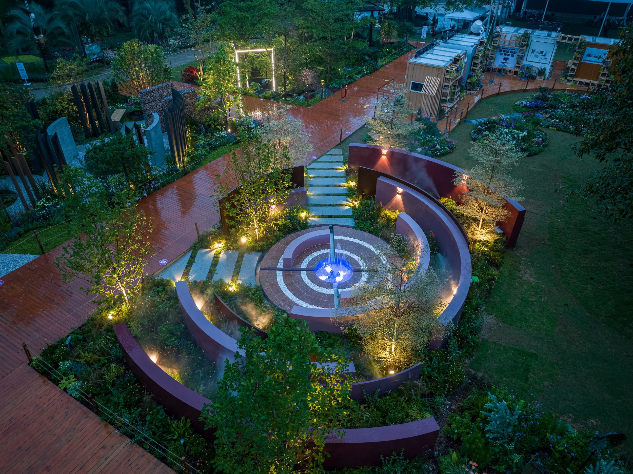Aerial view of a beautifully illuminated urban park at night featuring a circular water fountain, winding pathways, trees, landscaped gardens, and various greenery, with surrounding structures including light fixtures and display stands.