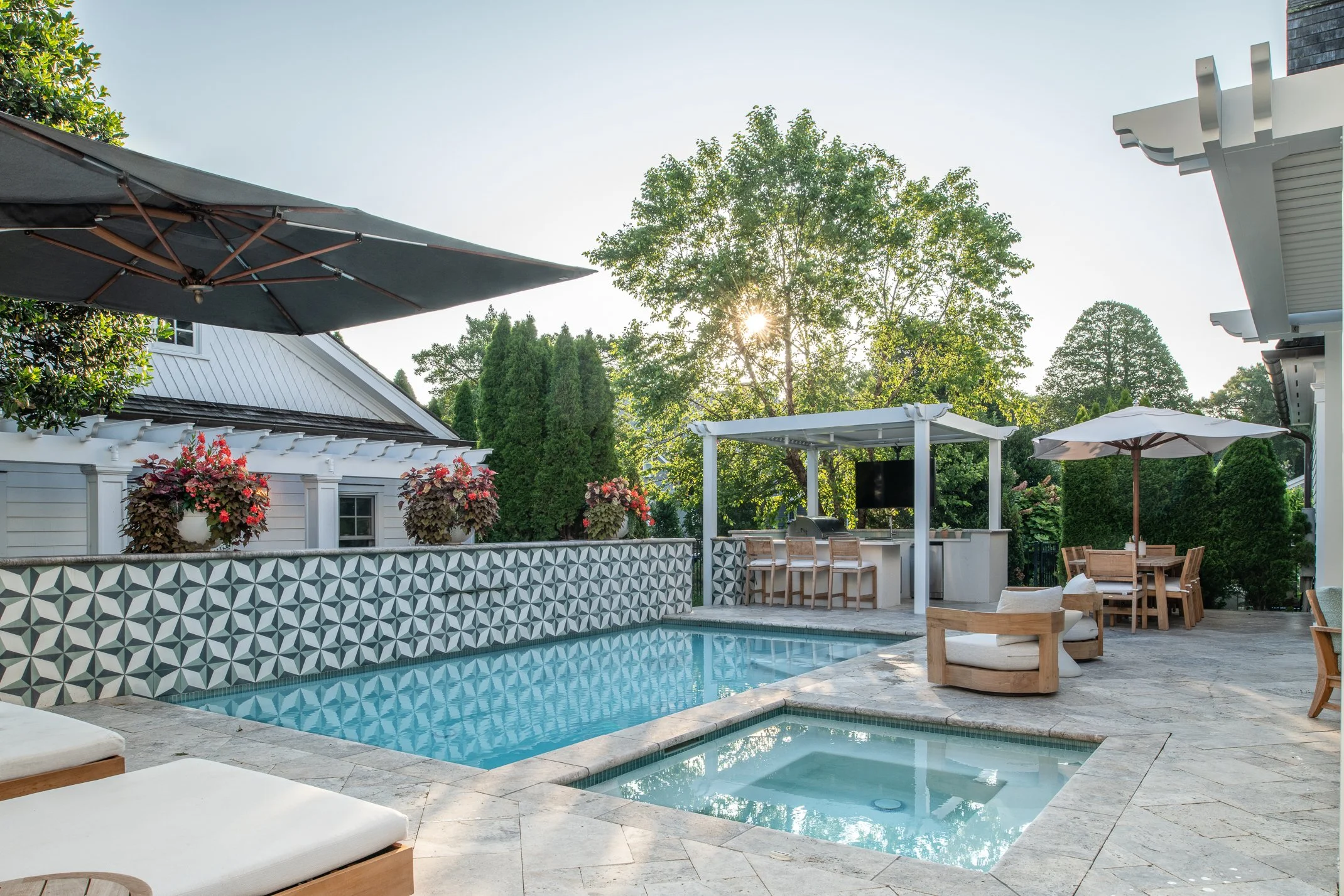 An outdoor backyard patio with a swimming pool, hot tub, shaded bar area, and seating, surrounded by greenery and flowering plants, during a sunny day.
