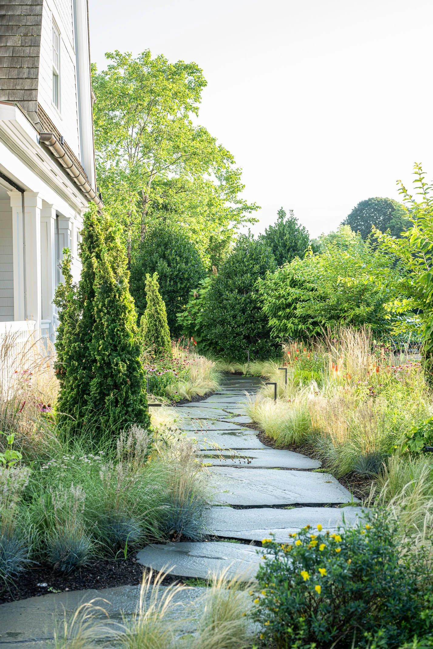 A garden pathway made of large stone slabs winding through lush greenery and colorful plants with a house on the left and tall trees in the background.