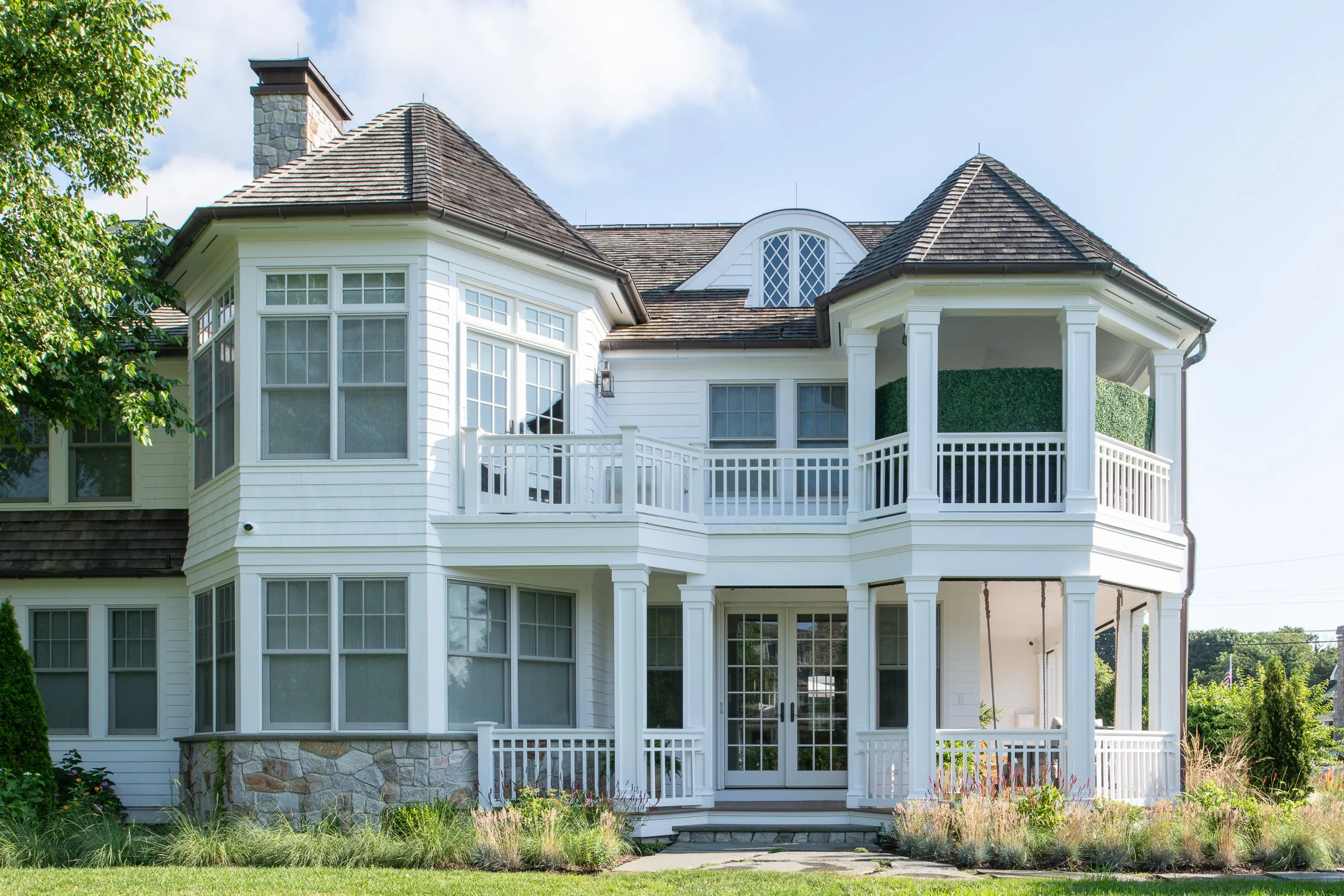 A large white two-story house with multiple balconies, bay windows, and a stone foundation, surrounded by a green lawn and trees.