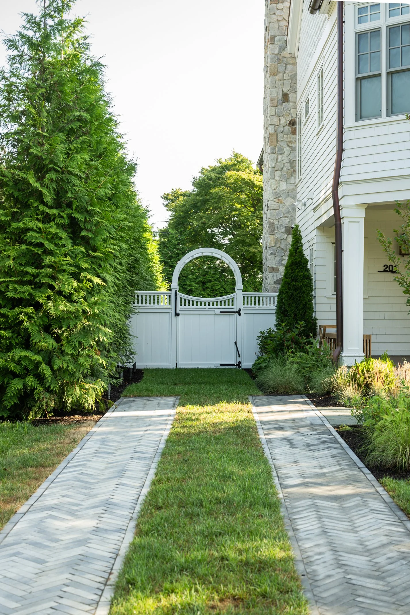 A white wooden gate with a curved top at the end of a brick pathway leading to a house, surrounded by green trees and plants.
