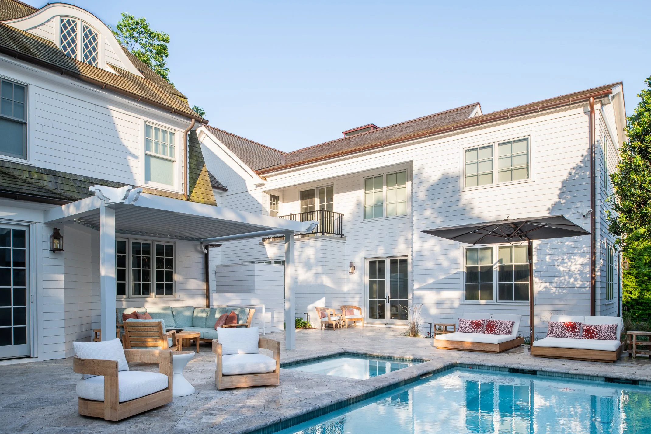 Backyard view of a white house with outdoor seating, a swimming pool, patio umbrellas, and trees, in bright sunlight.