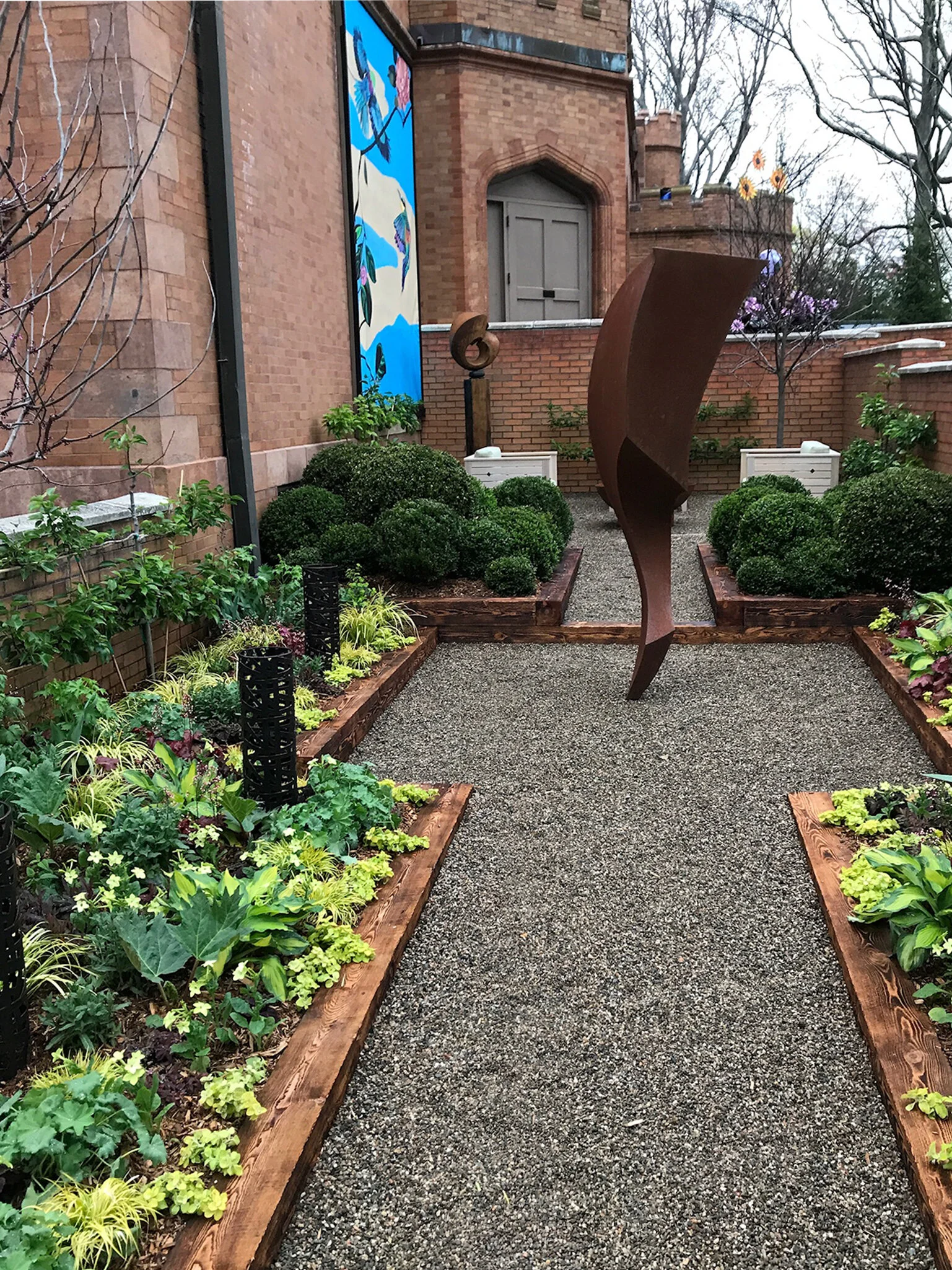 A small garden with raised flower beds containing various green plants and bushes, a gravel pathway, and abstract sculptures against a brick building with a mural painting.