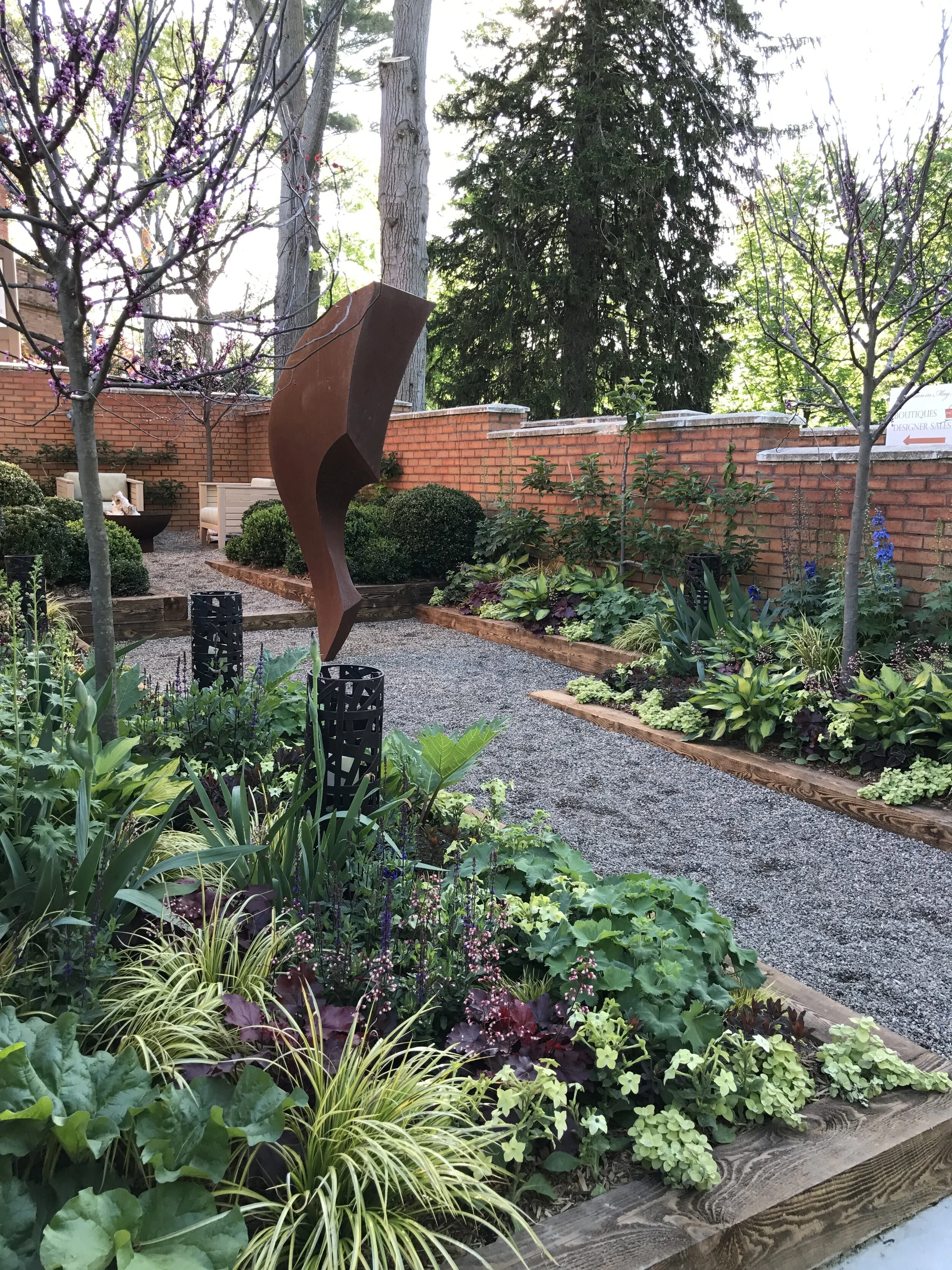 A small garden with a gravel path, various green plants, small trees, and purple flowering trees, surrounded by a brick wall. There are benches and a rust-colored abstract sculpture.