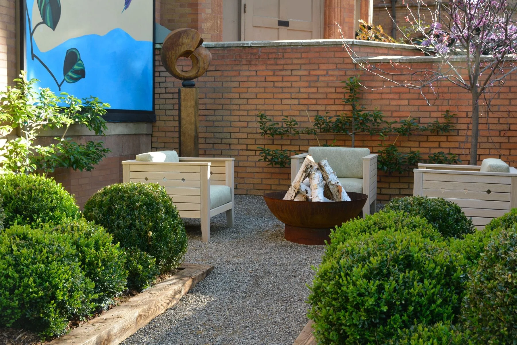 Outdoor patio with three white chairs surrounding a fire pit filled with split logs, green bushes, and trees with pink blossoms, enclosed by brick walls.