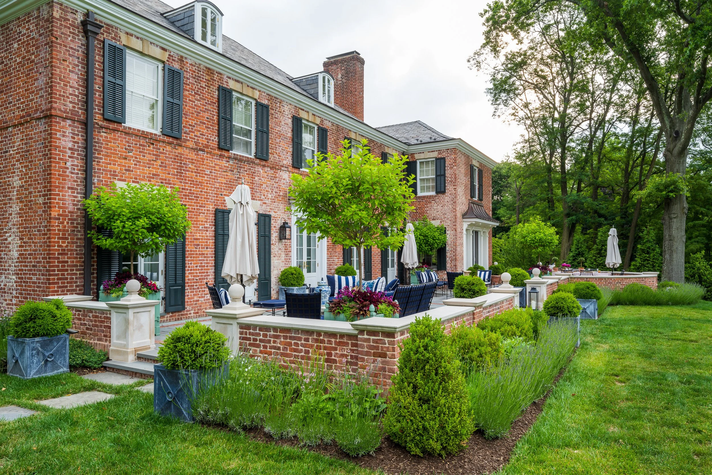 Large brick house with a spacious outdoor patio area decorated with potted plants, patio furniture, umbrellas, and lush green trees surrounding the property.