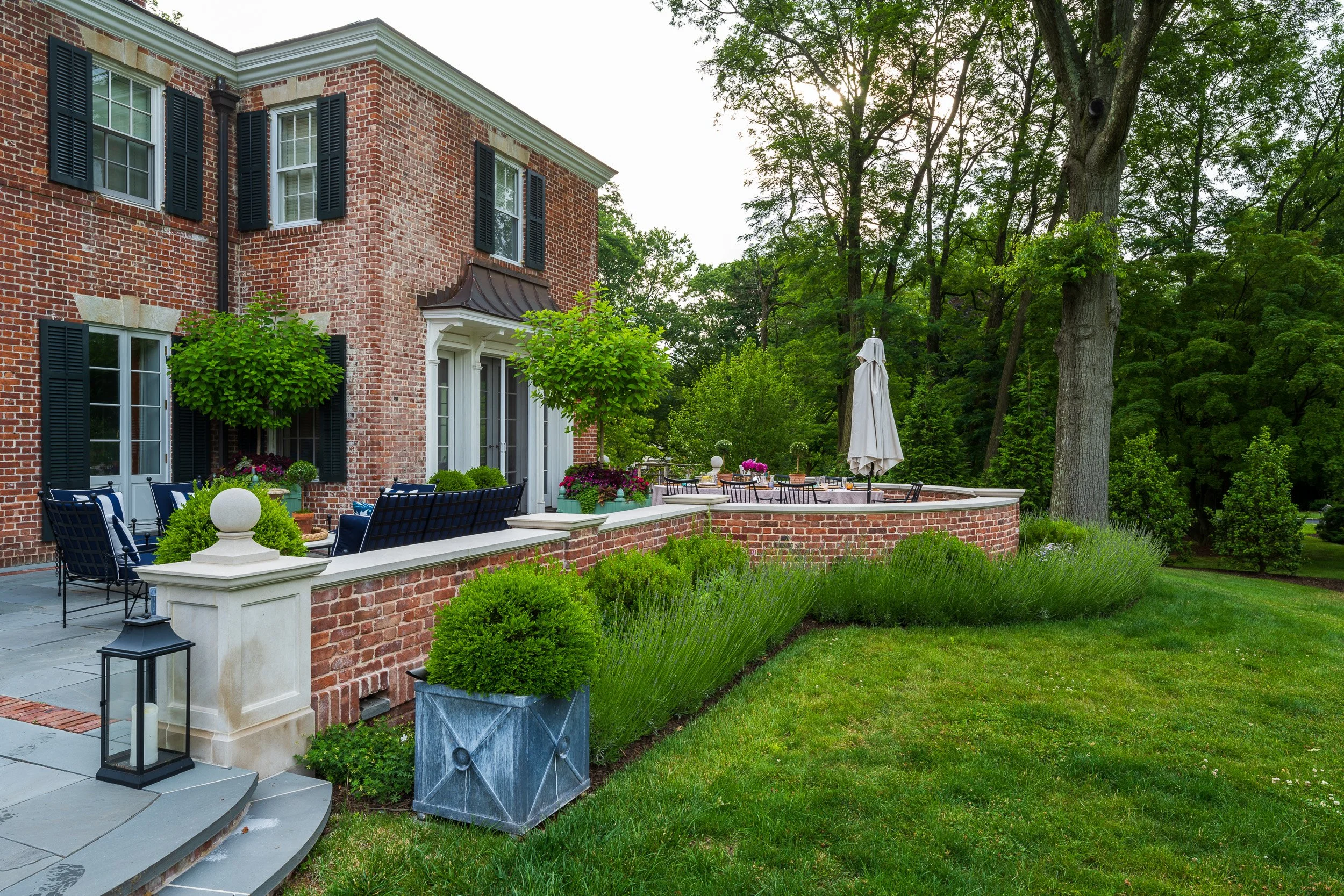 A backyard patio with outdoor furniture, potted plants, and a raised brick platform with a patio table and umbrella, surrounded by green trees and grass.
