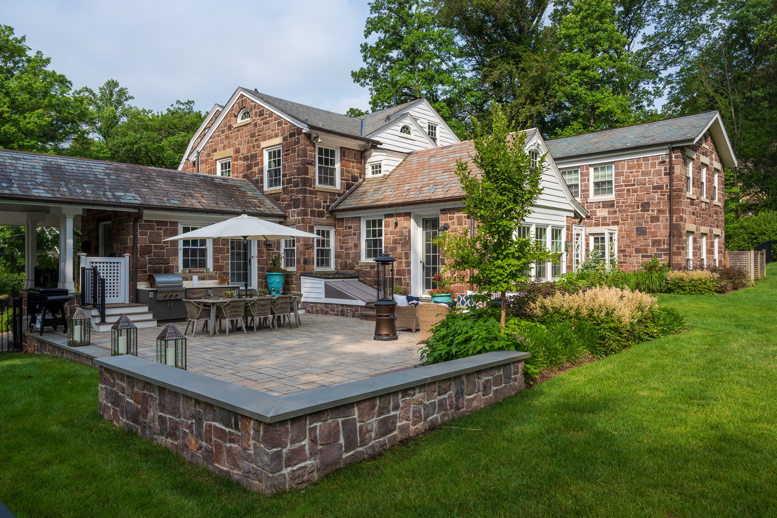 Elegant stone and white house with a courtyard patio, outdoor dining table, umbrella, and garden area surrounded by lush greenery and tall trees.