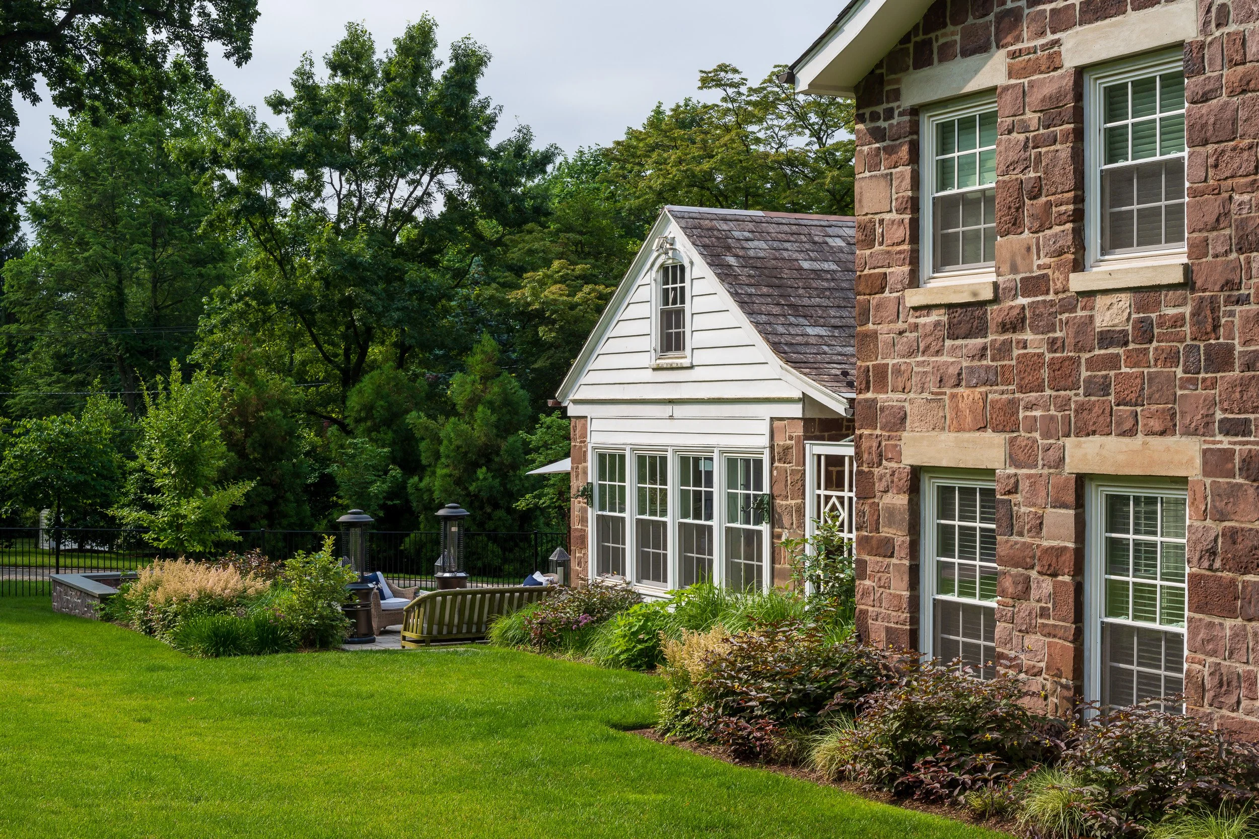 A backyard with a green lawn, outdoor seating, a stone and brick house, and lush trees in the background.