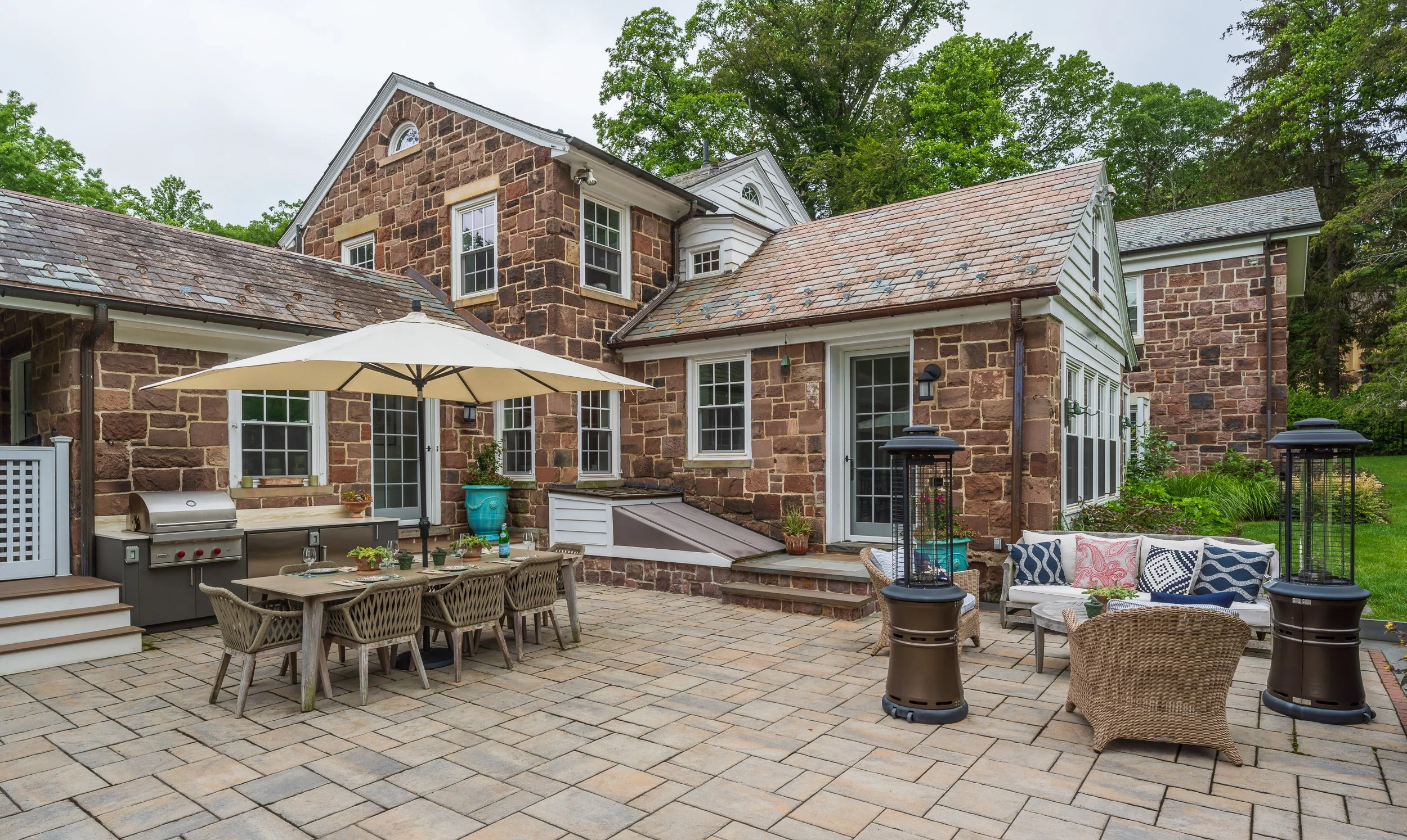 Backyard patio with outdoor furniture, a large umbrella, propane patio heaters, and a stone house with multiple levels and windows.