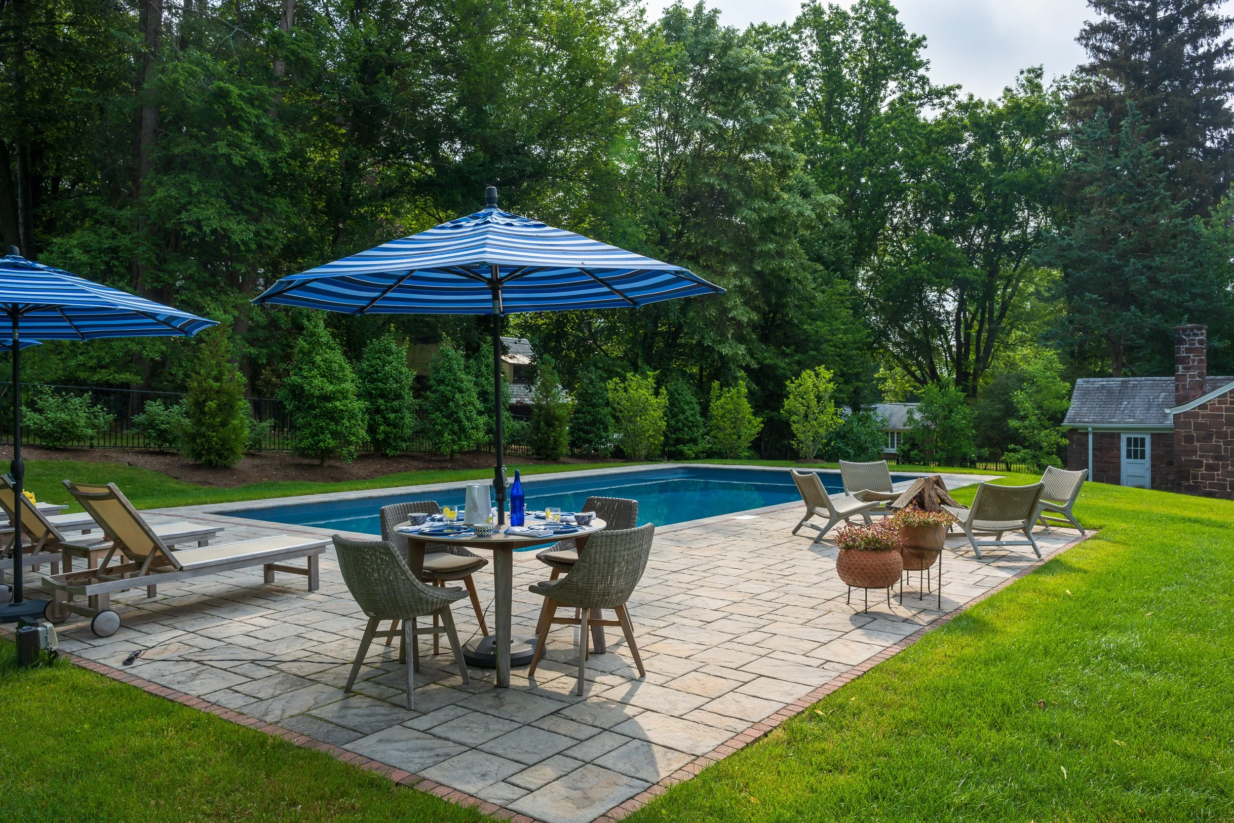 Backyard patio with a swimming pool, outdoor furniture including chairs and tables with umbrellas, and a lush green backdrop of trees and grass.