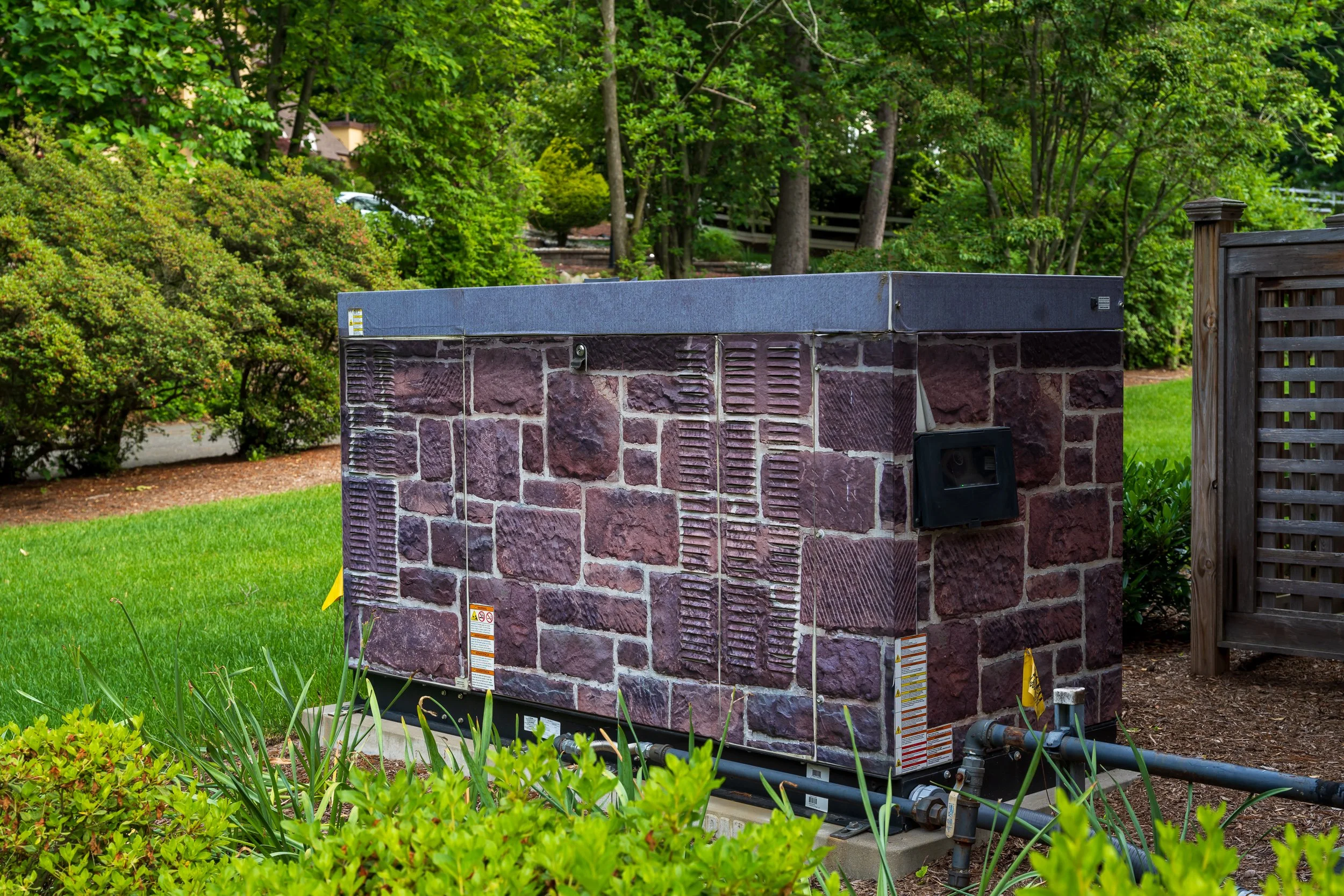 A utility box with a stone brick pattern surrounded by green grass and bushes in a park or garden area.