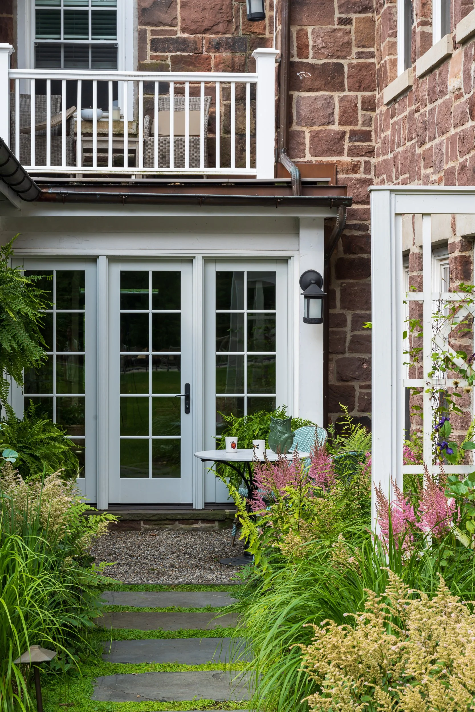 A garden patio with outdoor furniture, potted plants, and flowering plants surrounding a gravel and stone pathway leading to glass doors and a balcony above. The setting is part of a brick house.