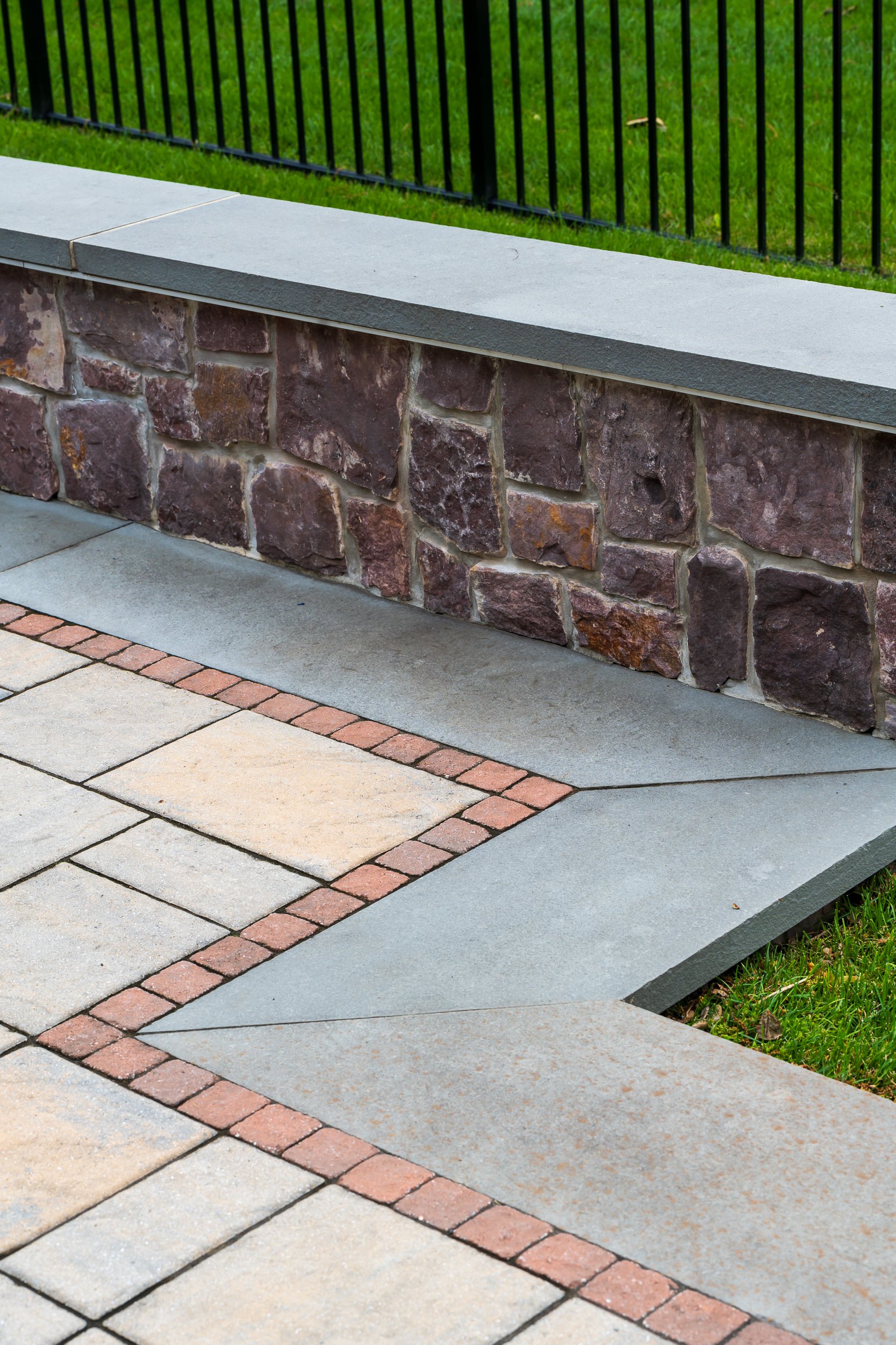 Close-up of a stone and concrete patio with a low stone wall and a black metal railing on a grassy area.