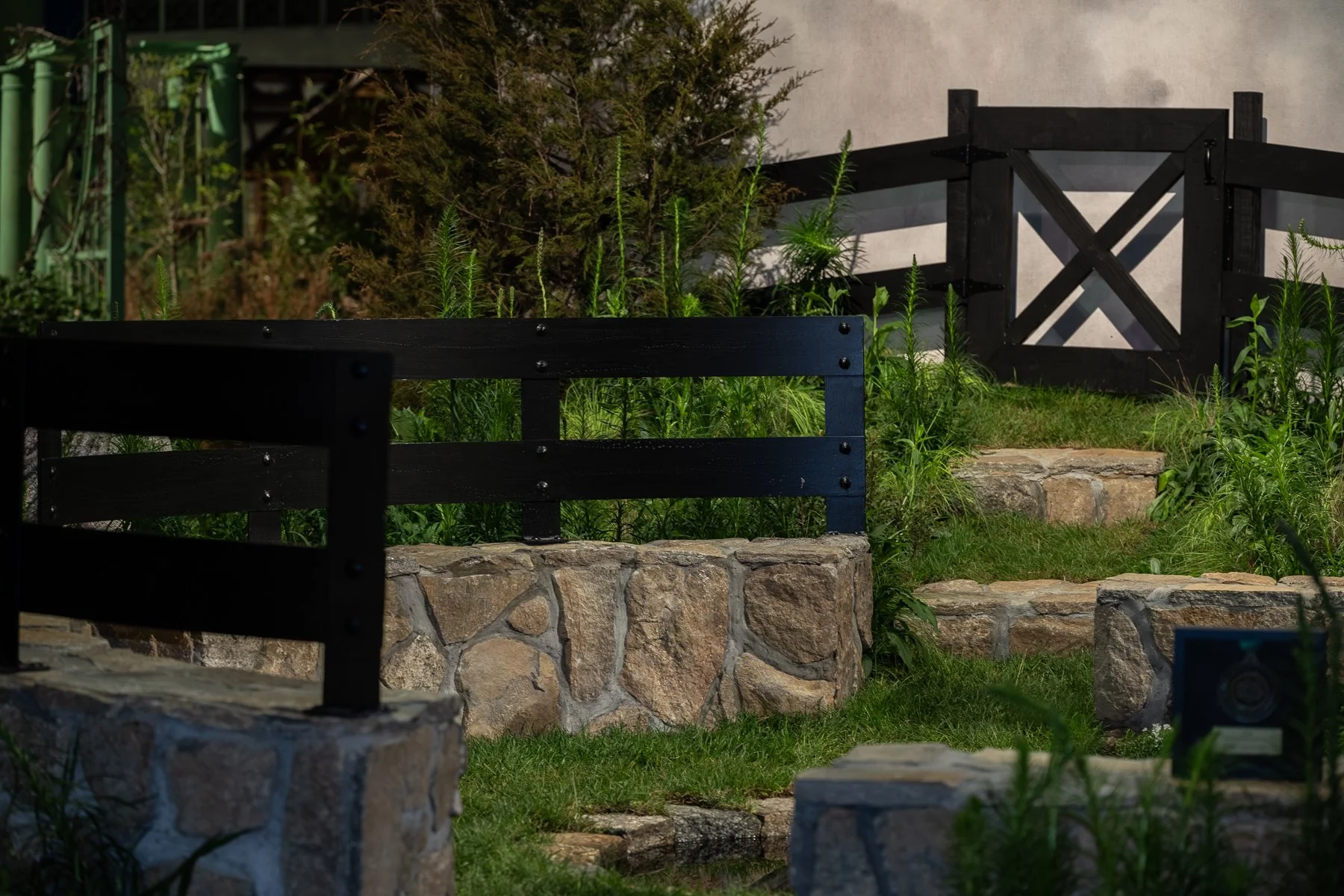 A garden scene with stone steps, black wooden benches, green plants, and a beige wall, with some greenery in the background.