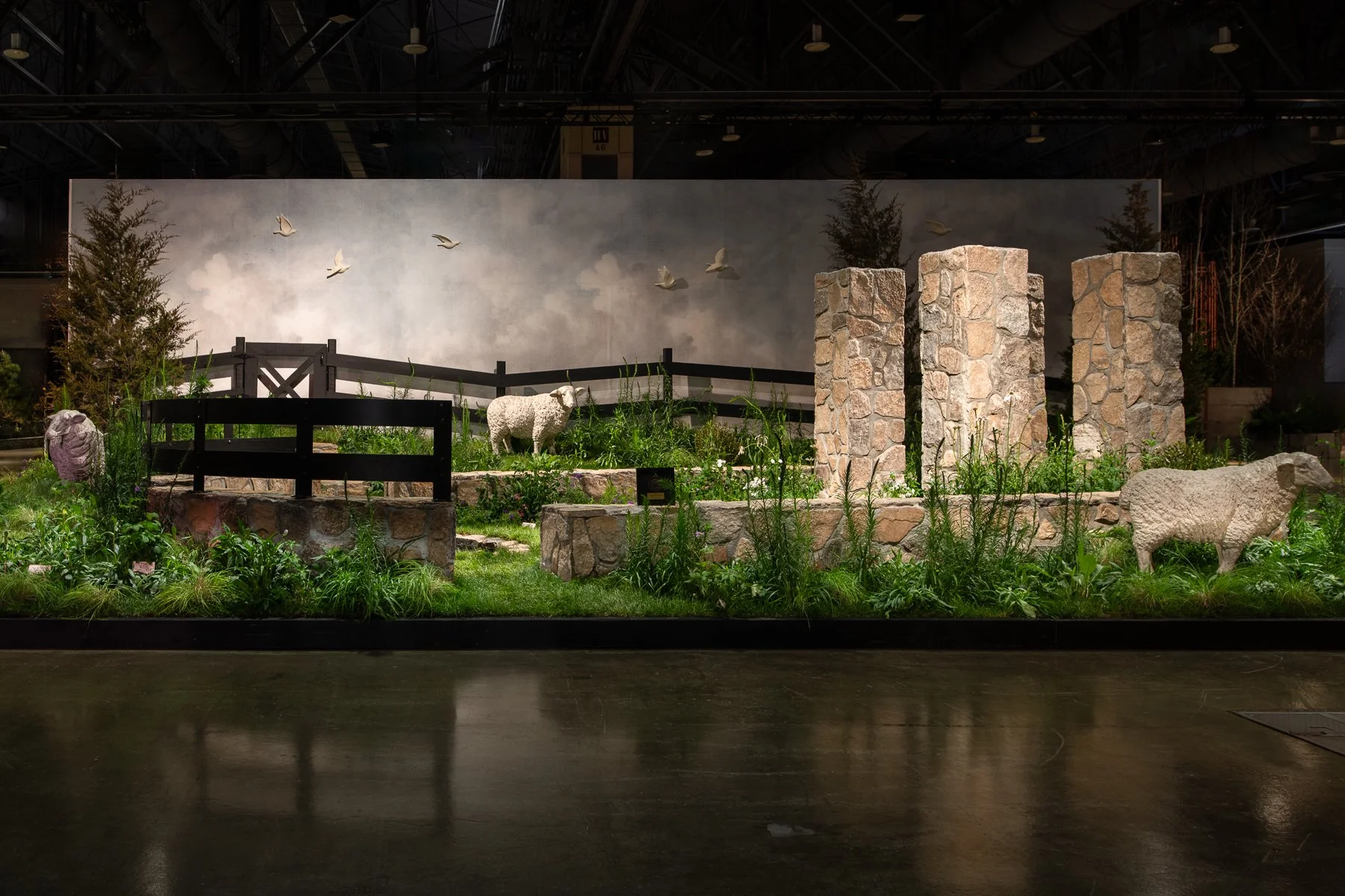An indoor diorama of a farm scene with stone pillars, animal statues of a sheep and a ram, greenery, a small wooden fence, and a backdrop painting of a cloudy sky with flying birds.