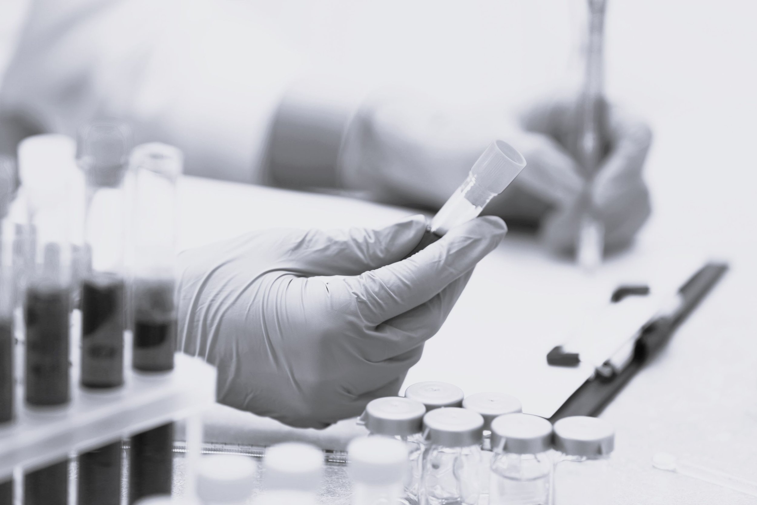 Person wearing gloves handling a test tube filled with liquid in a laboratory setting, with test tubes and containers in the background.