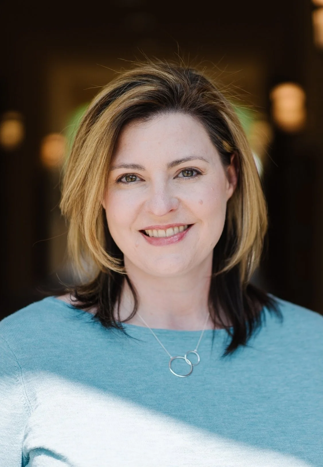 Stephanie Vance, Licensed Professional Counselor in Flowood, MS, with shoulder-length, light brown hair, smiling at the camera, wearing a light blue top and a silver necklace with two linked rings, blurred warm background.