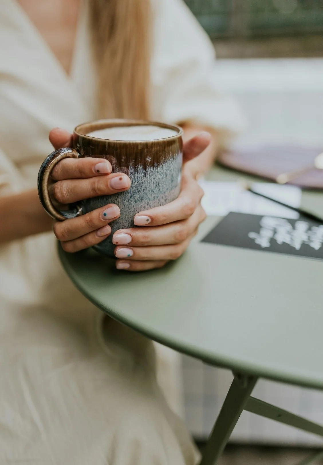 Person holding a large ceramic mug of coffee or tea with both hands, sitting at a green table near a window.