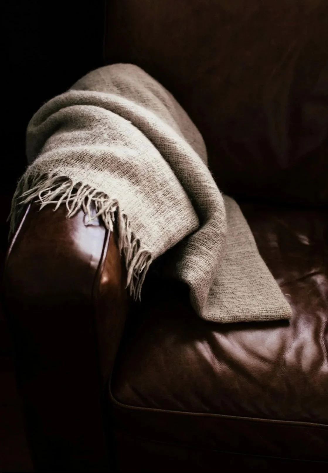 Lying on a brown leather couch, a pair of crossed legs with beige pants, a beige woven blanket with fringes, and a brown leather shoe.