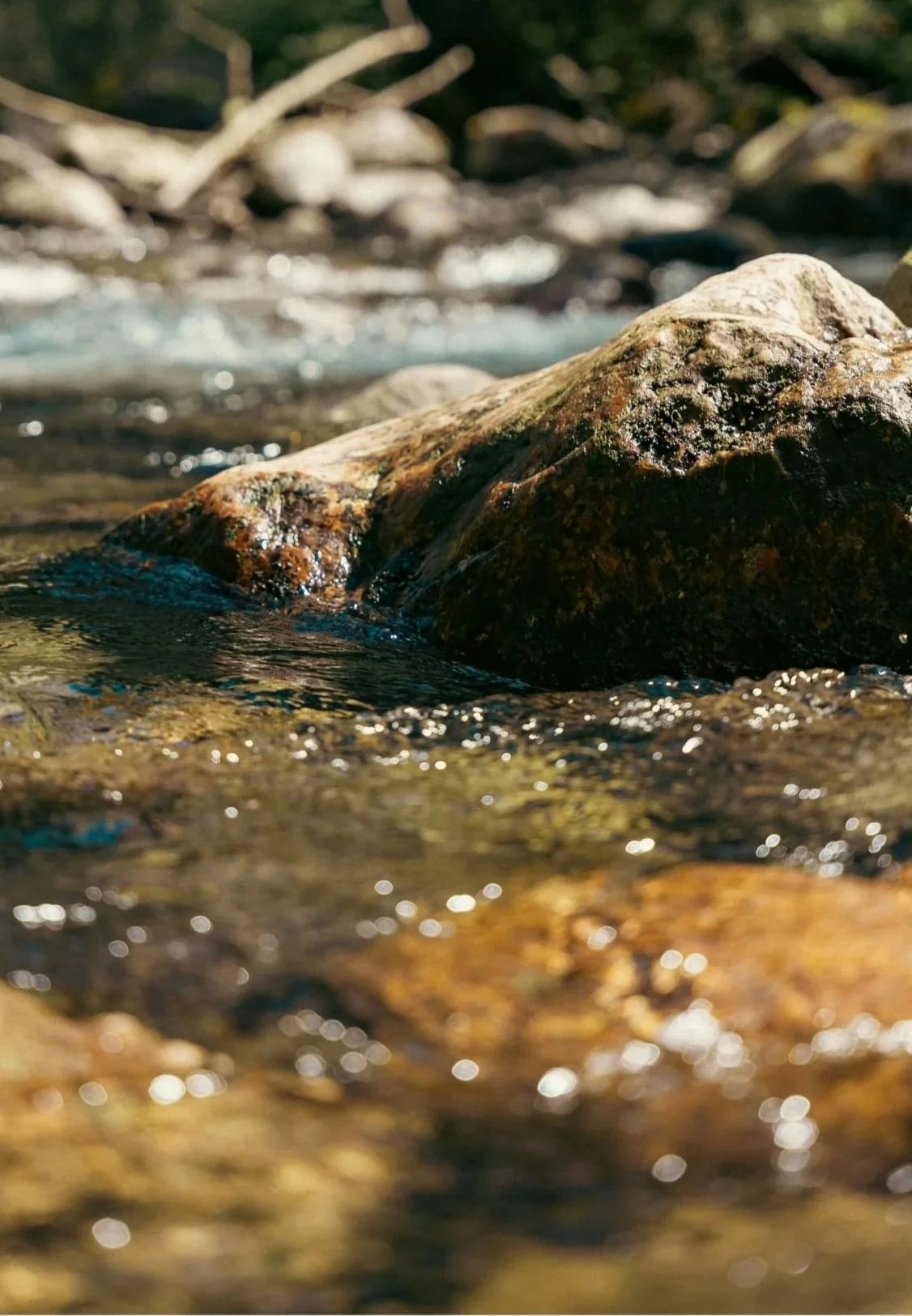 Close-up of a large rock in a flowing stream, with water moving around it and small pebbles visible in the stream bed, outdoors during daytime.