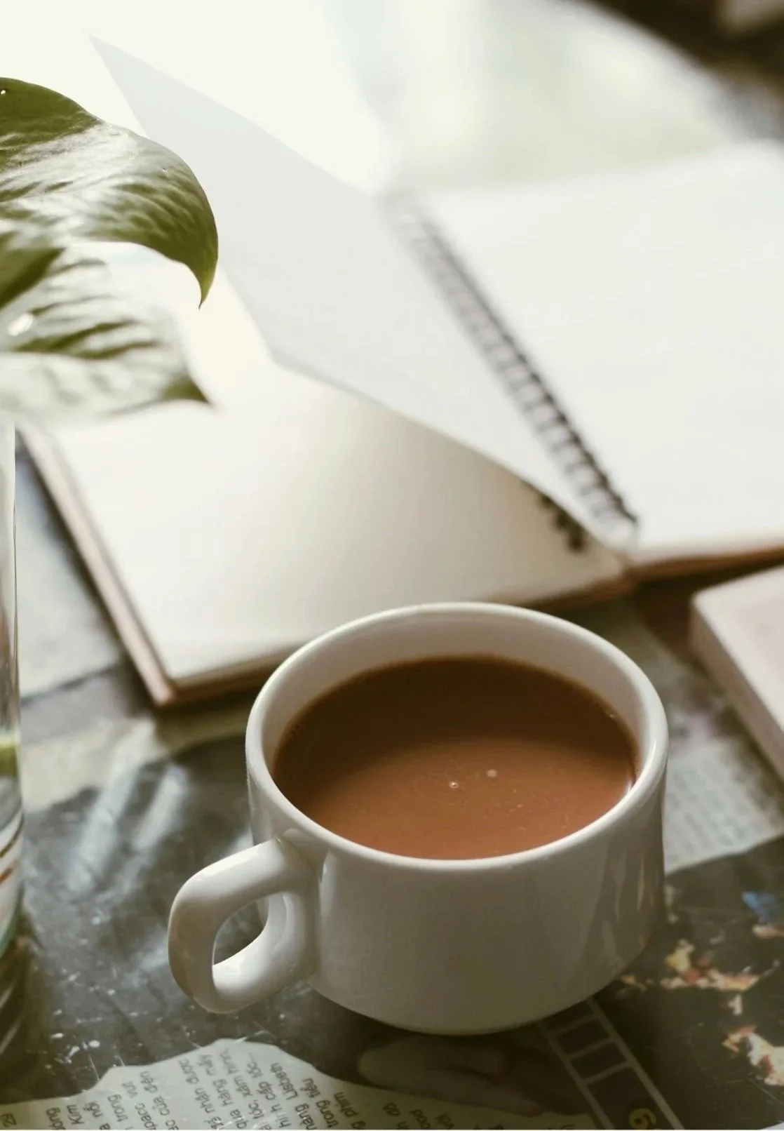 A white mug filled with coffee on a table, an open book in the background, a potted plant with large green leaves partially visible on the side, and a piece of paper with printed text in the foreground.