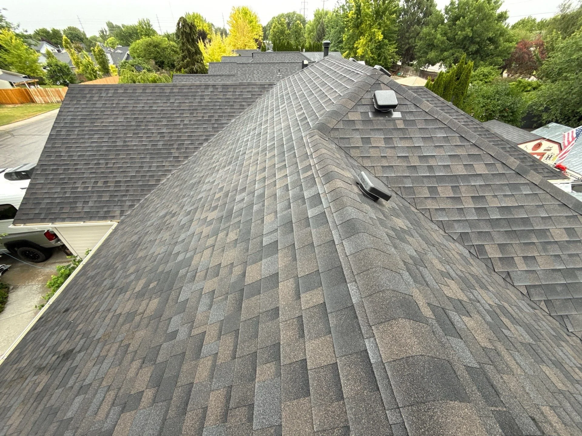 An aerial view of multiple rooftops with asphalt shingles, surrounded by trees and a neighborhood with houses and drives.