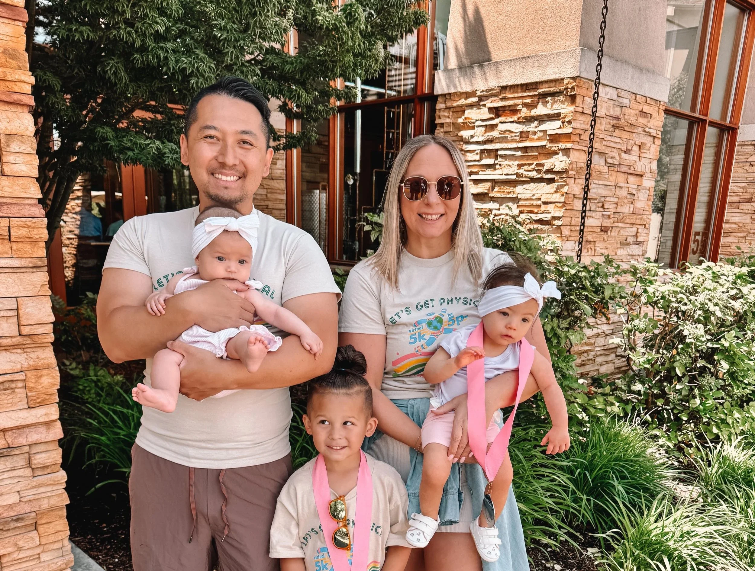 A diverse family of six, including two adults and four children, smiling outdoors in front of a brick building with large windows, green trees, and plants.