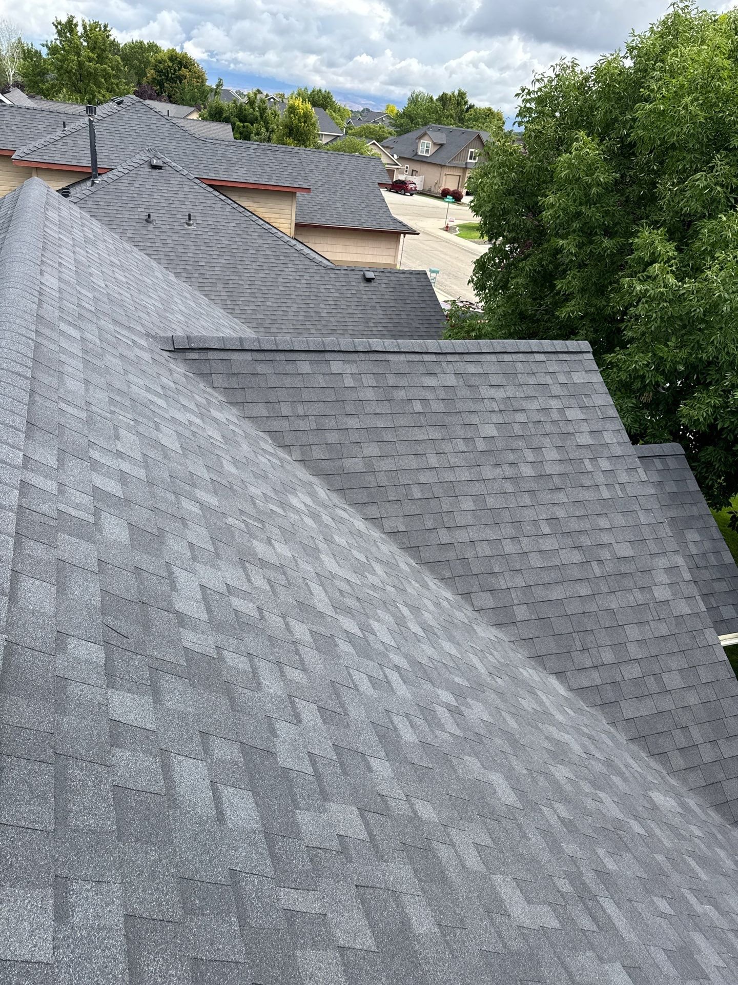 Multiple rooftops with gray shingles, trees, houses, and a cloudy sky in the background.