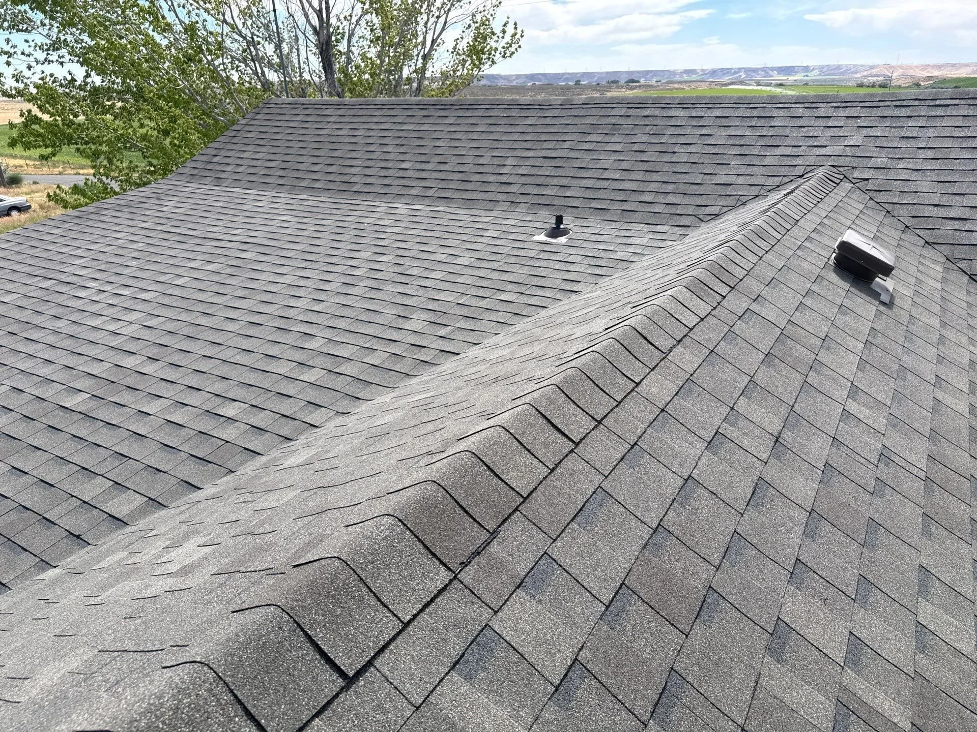 View of a residential roof with gray asphalt shingles, two vent pipes, and a ridge vent, with a tree and distant hills in the background.