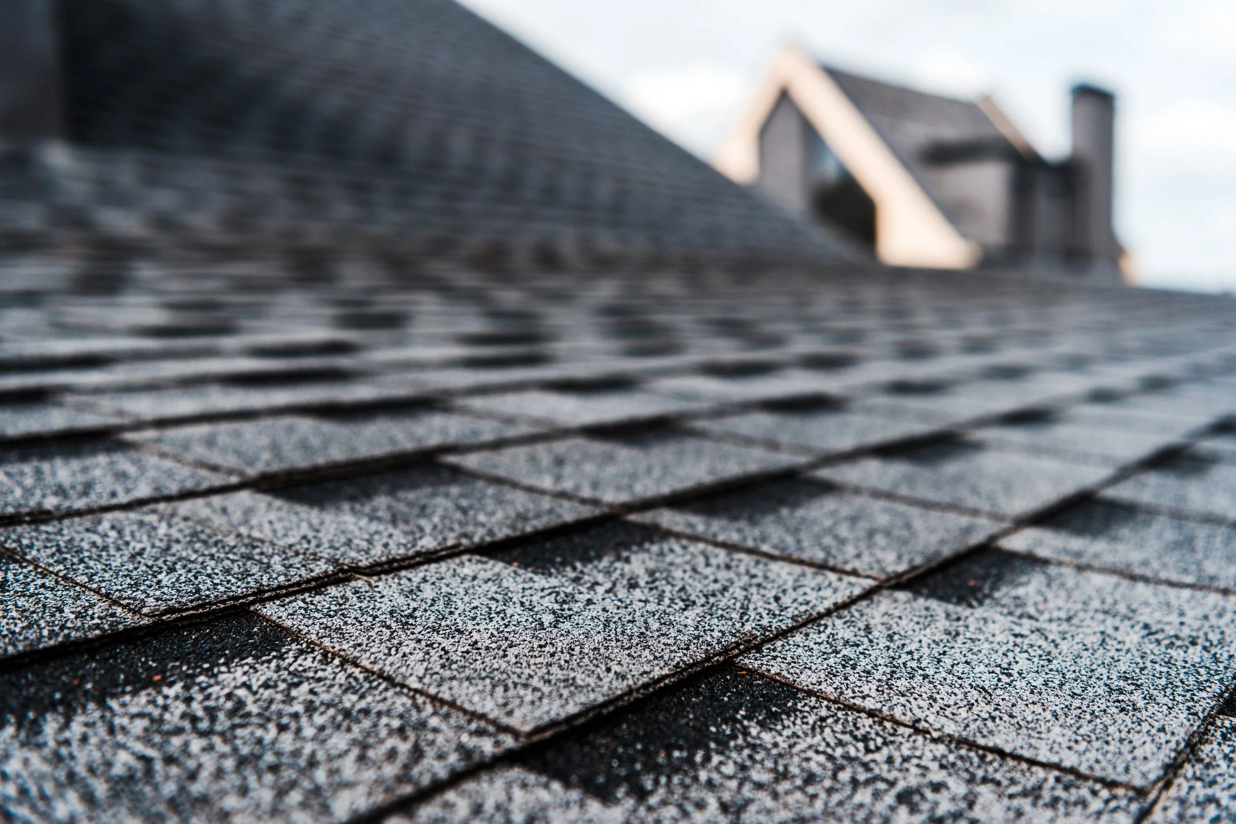 Close-up of a textured, gray asphalt shingle roof with a blurred background of a house and sky.