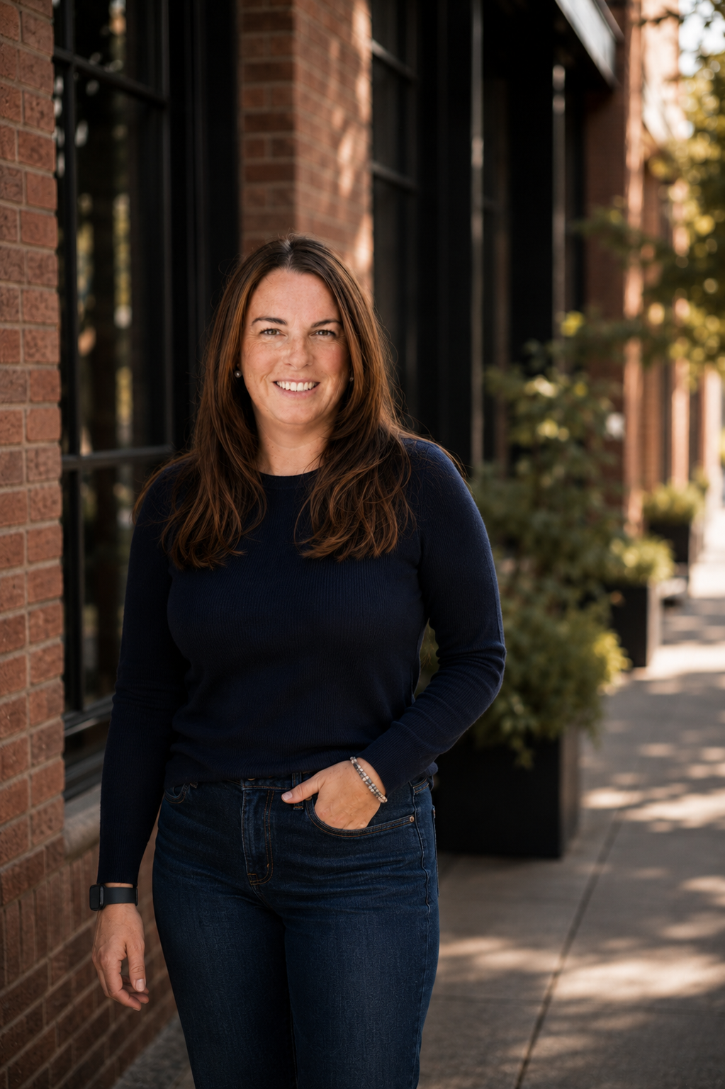 A woman with brown hair and a navy blue sweater standing outside on a sidewalk with brick buildings and trees in the background.