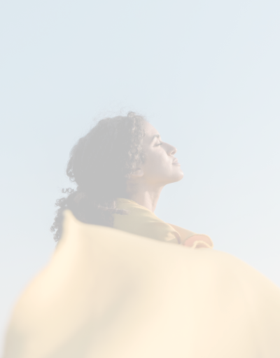 A woman with curly hair standing outdoors with her eyes closed, facing the sky, holding a large yellow fabric or garment.